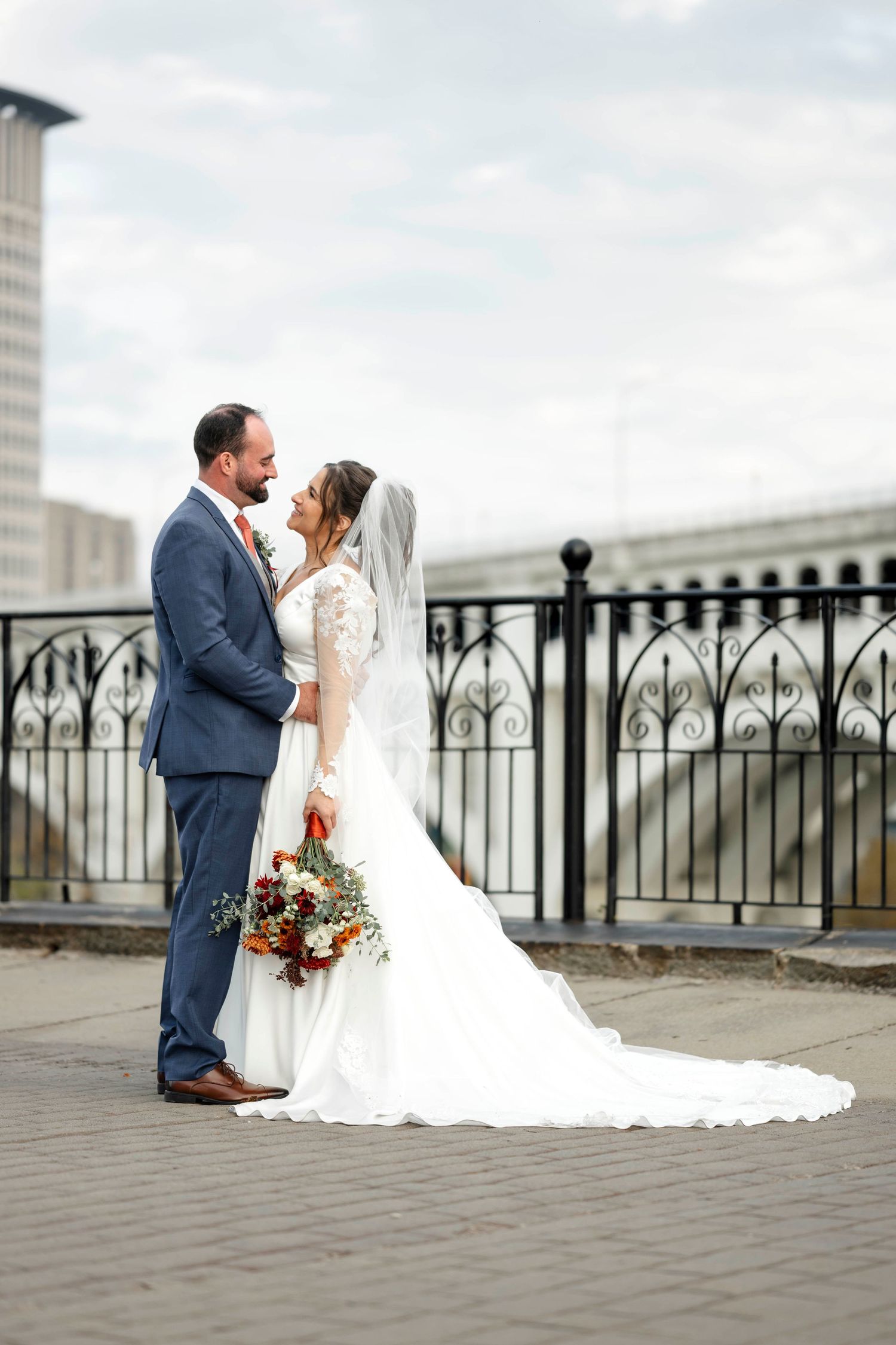 A couple in wedding attire shares romantic moments on a balcony with ornate railings and city views in the background.