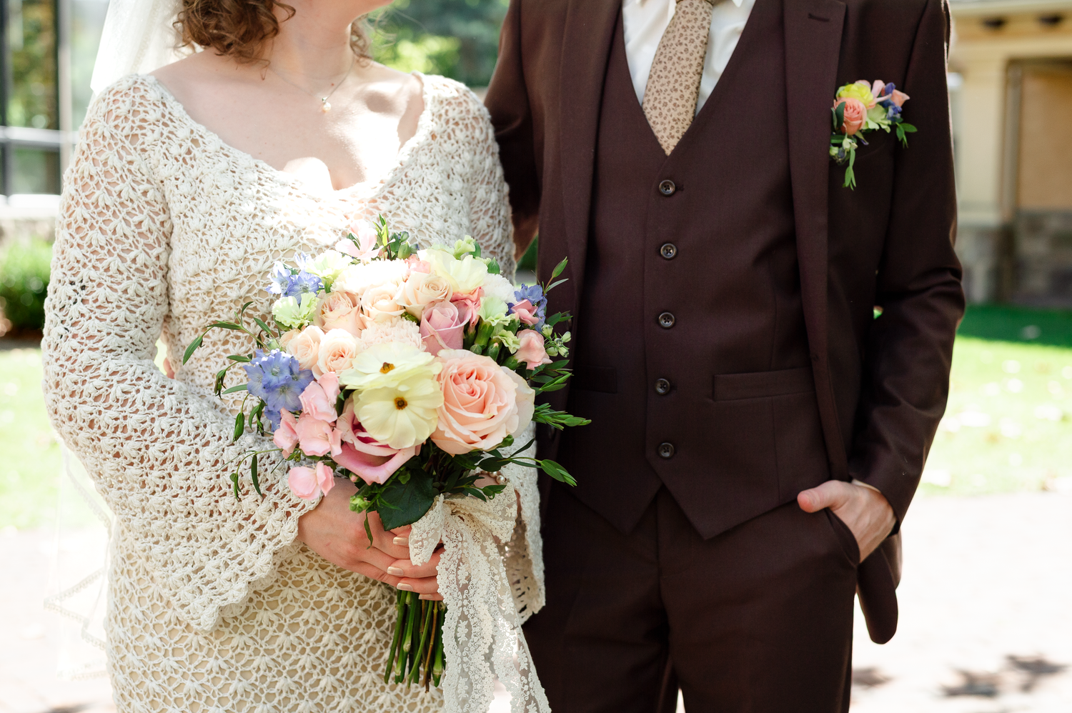 Close up of wedding couple with pastel bouquet, bride in lace dress and groom in brown suit.