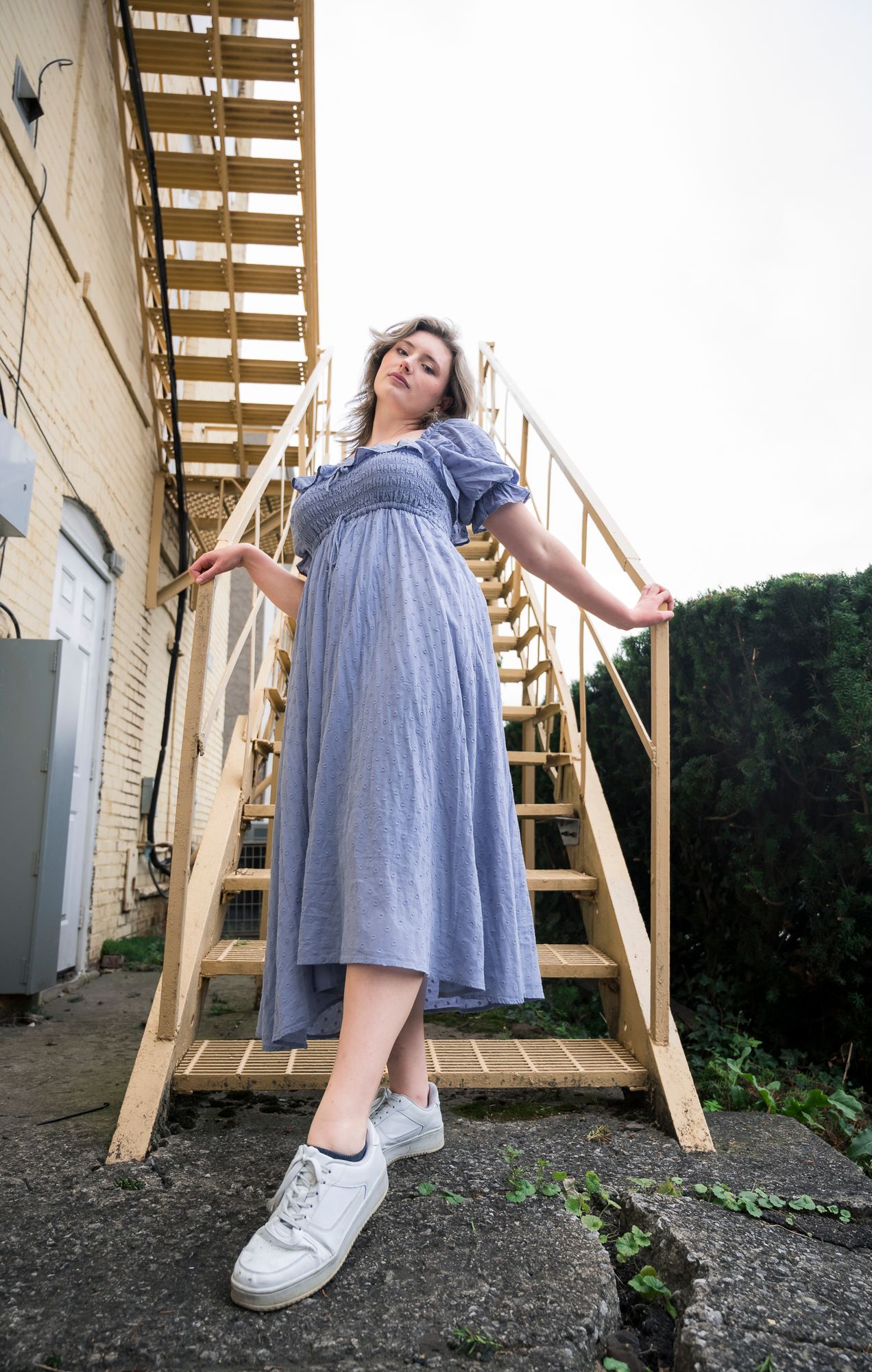 Person in a blue dress poses playfully on wooden stairs outdoors.