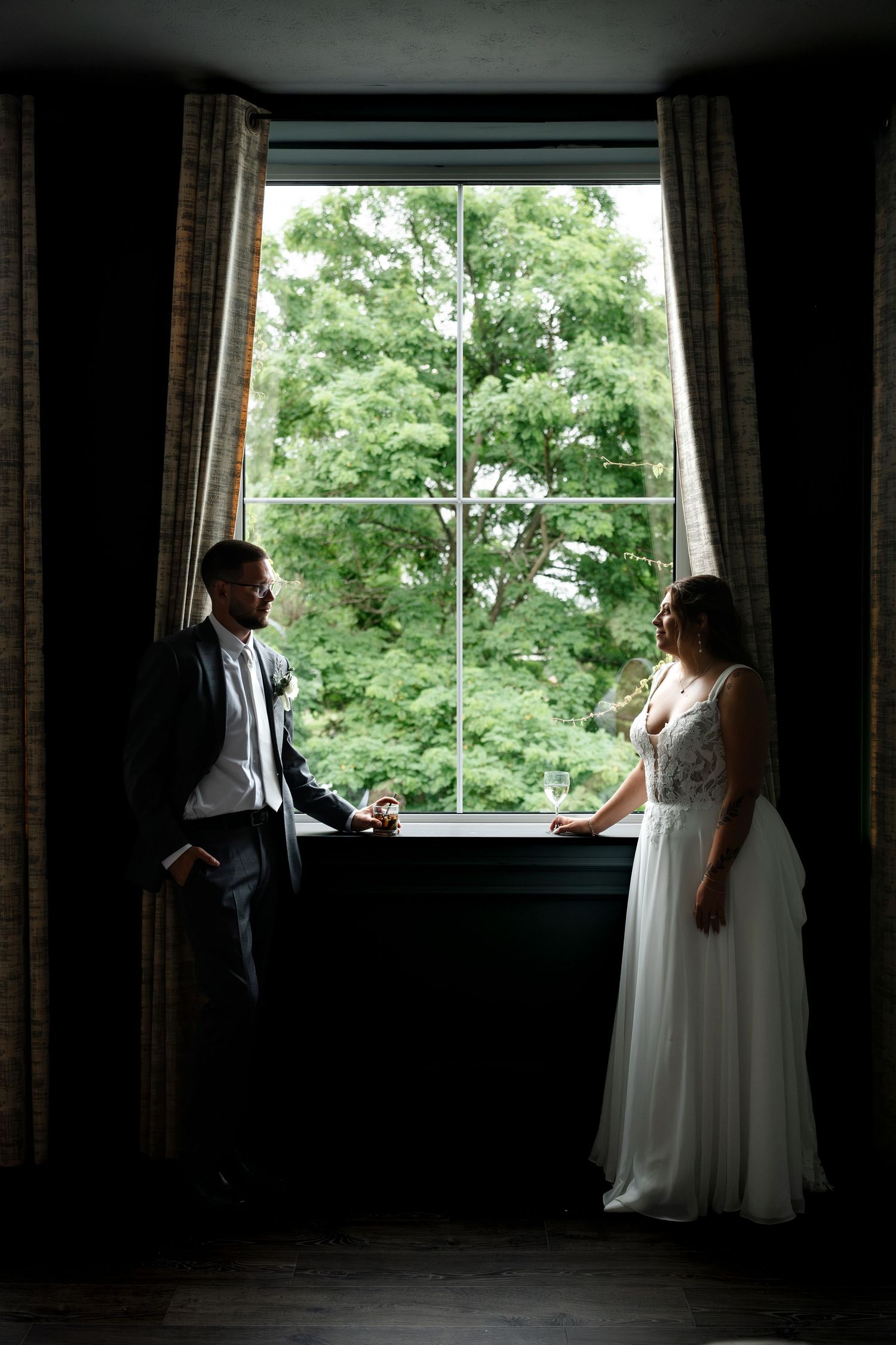Bride and groom holding hands by a large window with lush green trees, silhouetted in soft natural light.