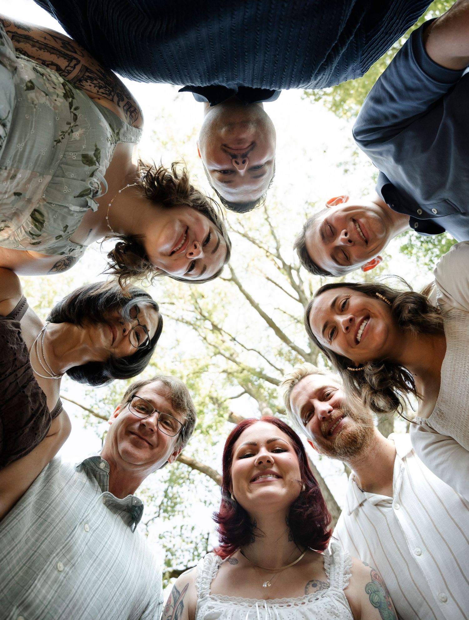 Group of friends forming a circle huddle photographed from below, smiling faces surrounded by trees.