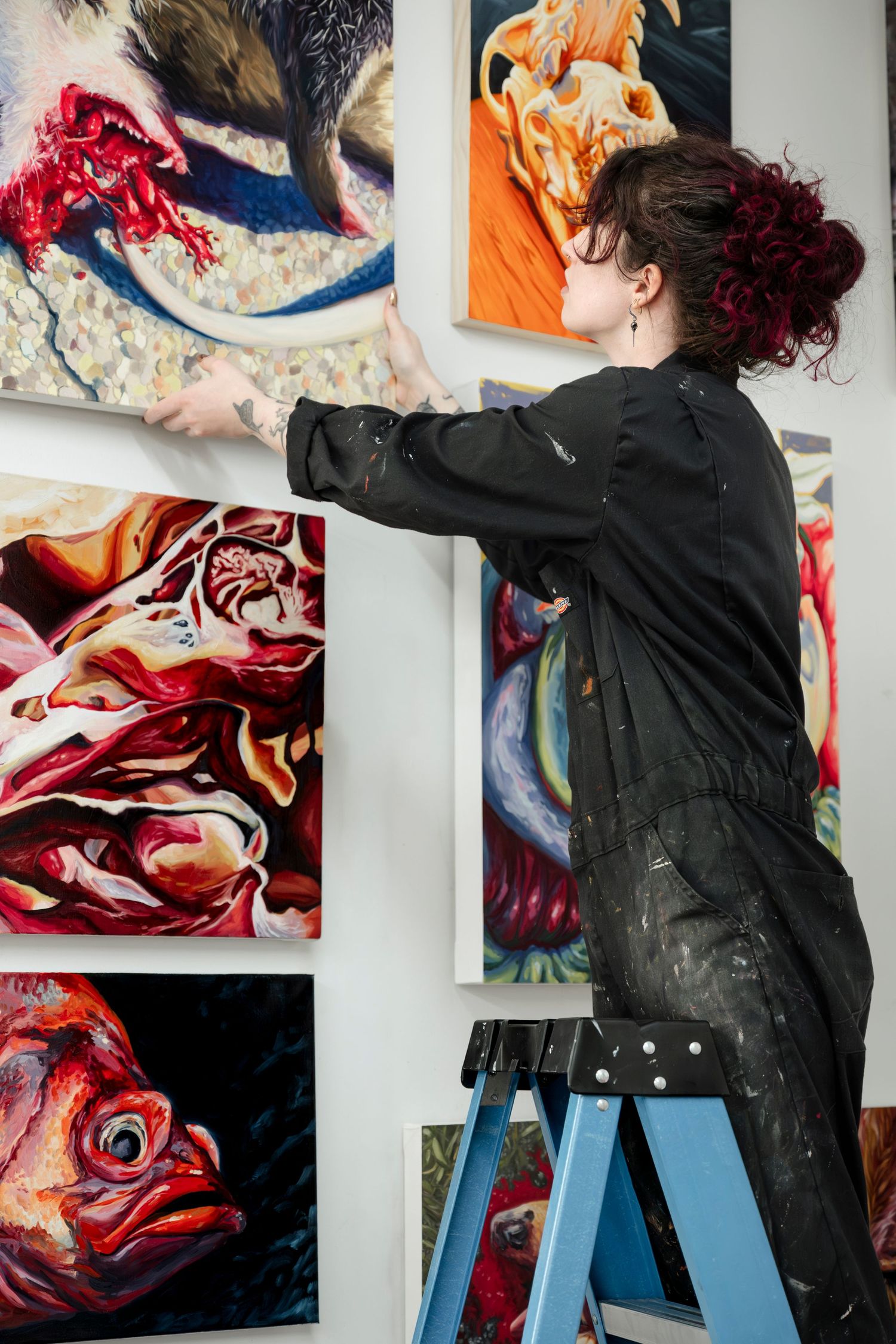 Artist in black jacket works on vibrant abstract paintings displayed on white gallery walls.