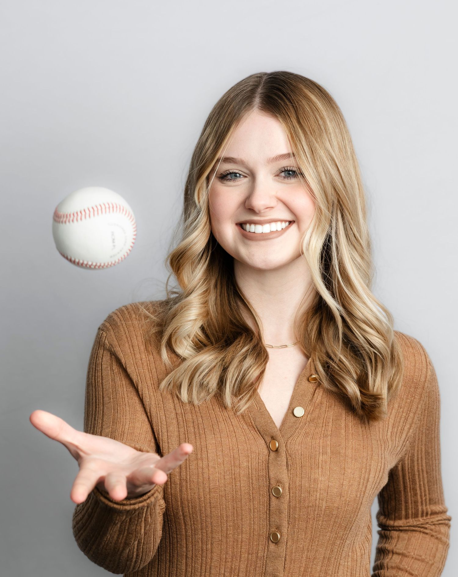 Playful portrait series showing someone in brown sweater tossing a baseball against gray backdrop.