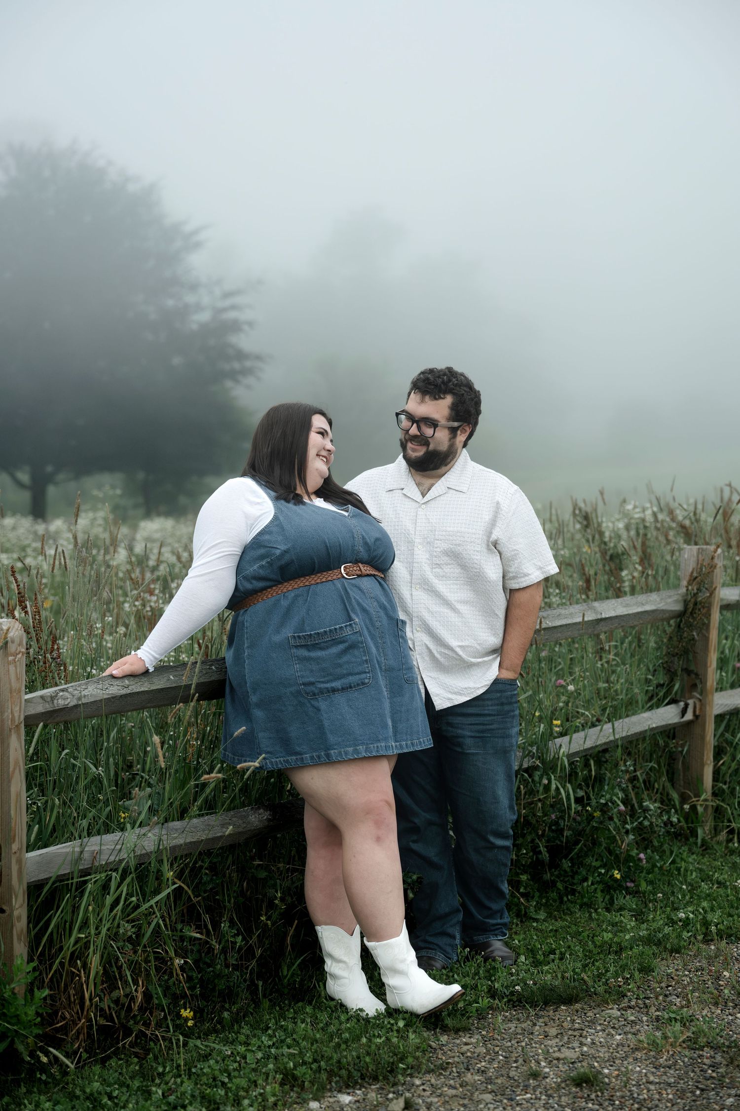 A couple smiles at each other leaning on a wooden fence in a foggy field, woman in denim dress and white boots.