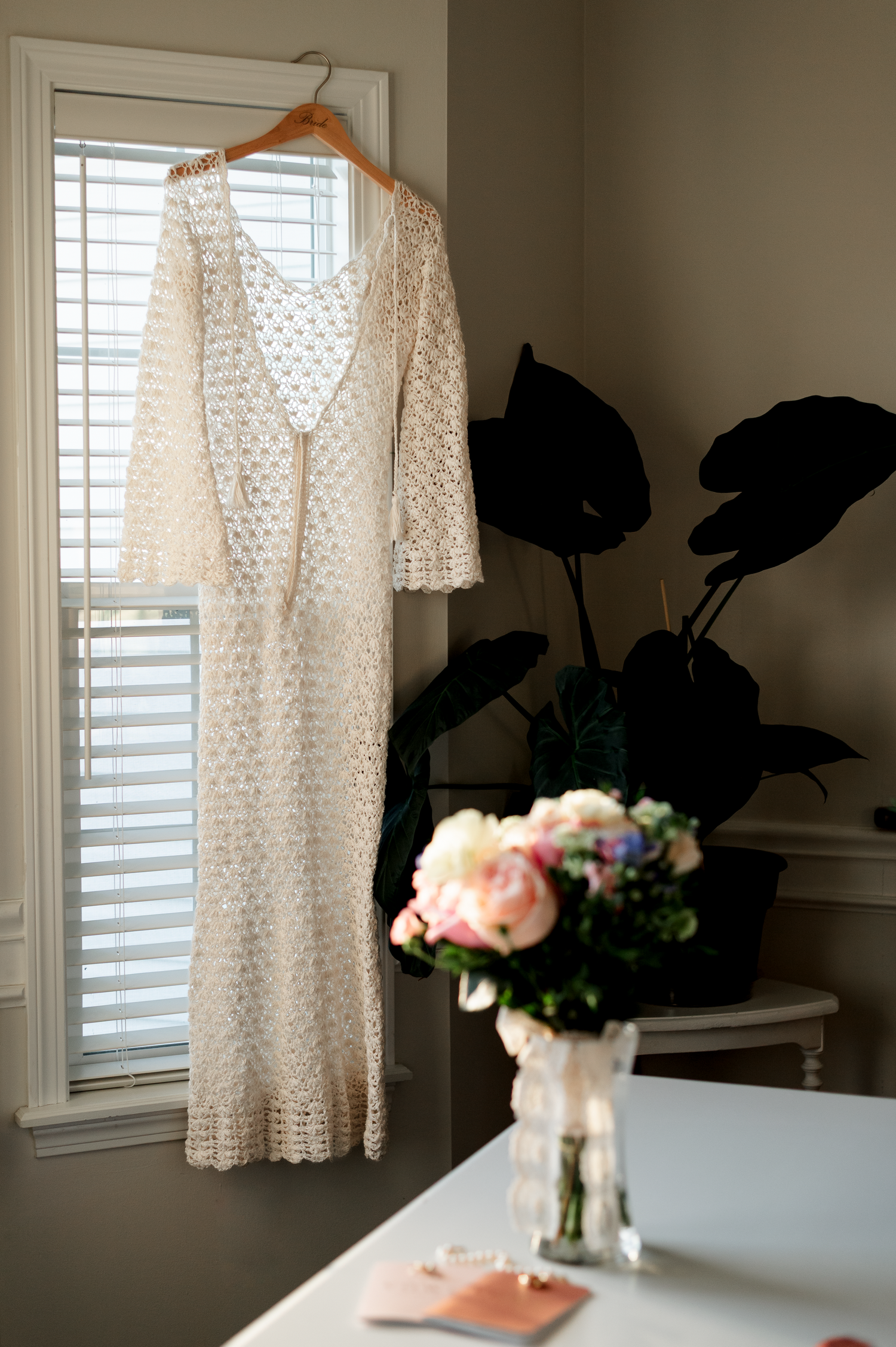 White lace wedding dress hanging by window with pink roses in vase.