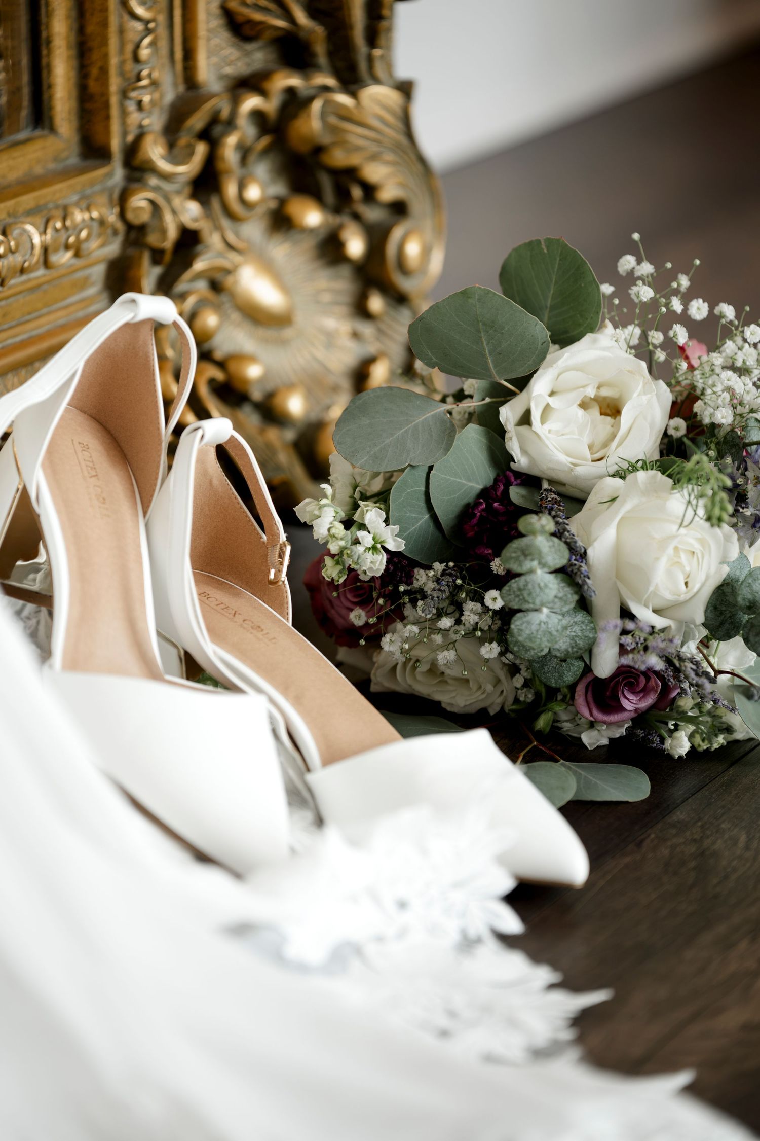 White bridal heels and elegant bouquet with roses, eucalyptus, and succulents beside a ornate gold mirror.
