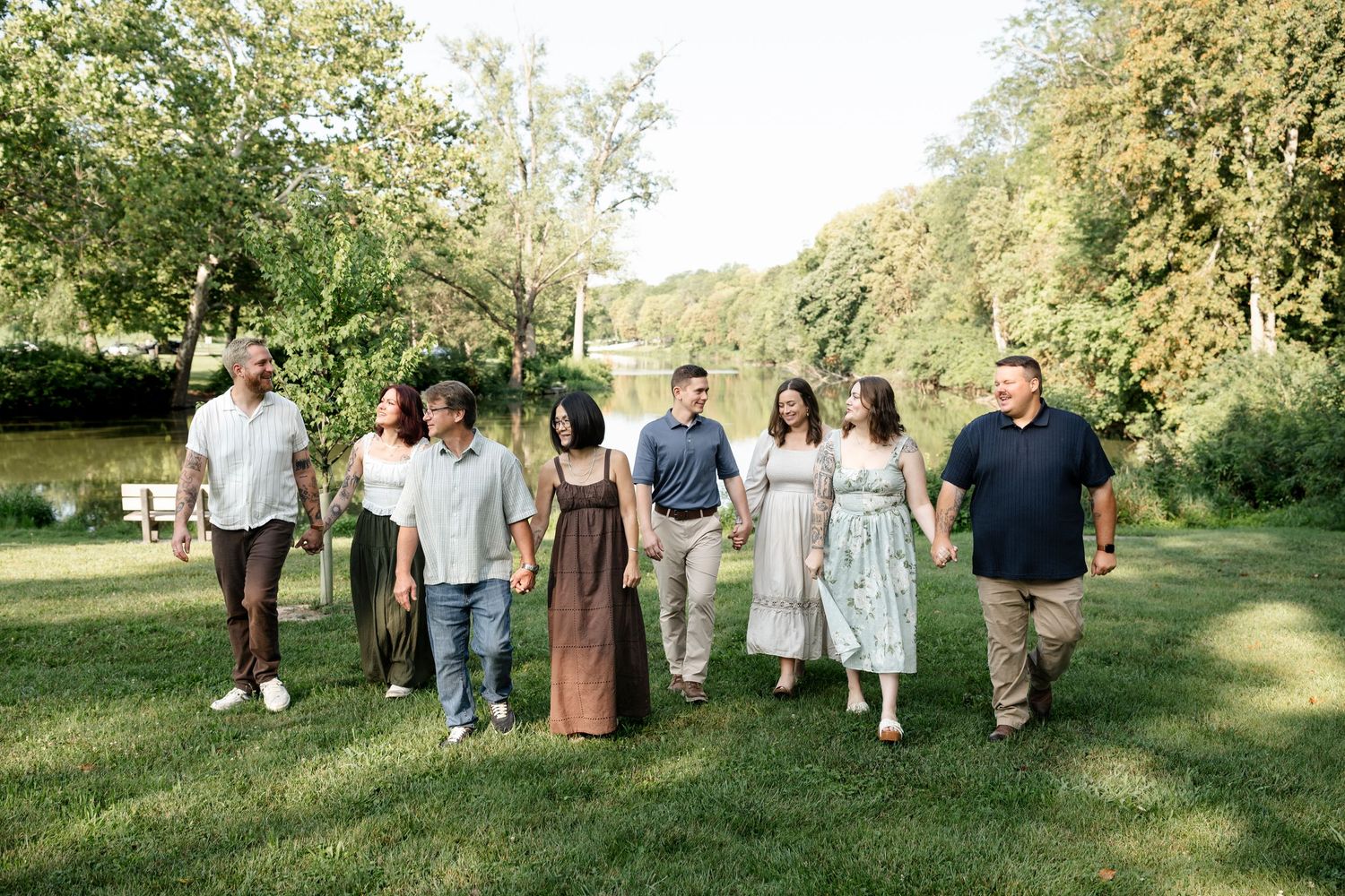 A large family group walks together on green grass in a park with trees and a pond in the background.