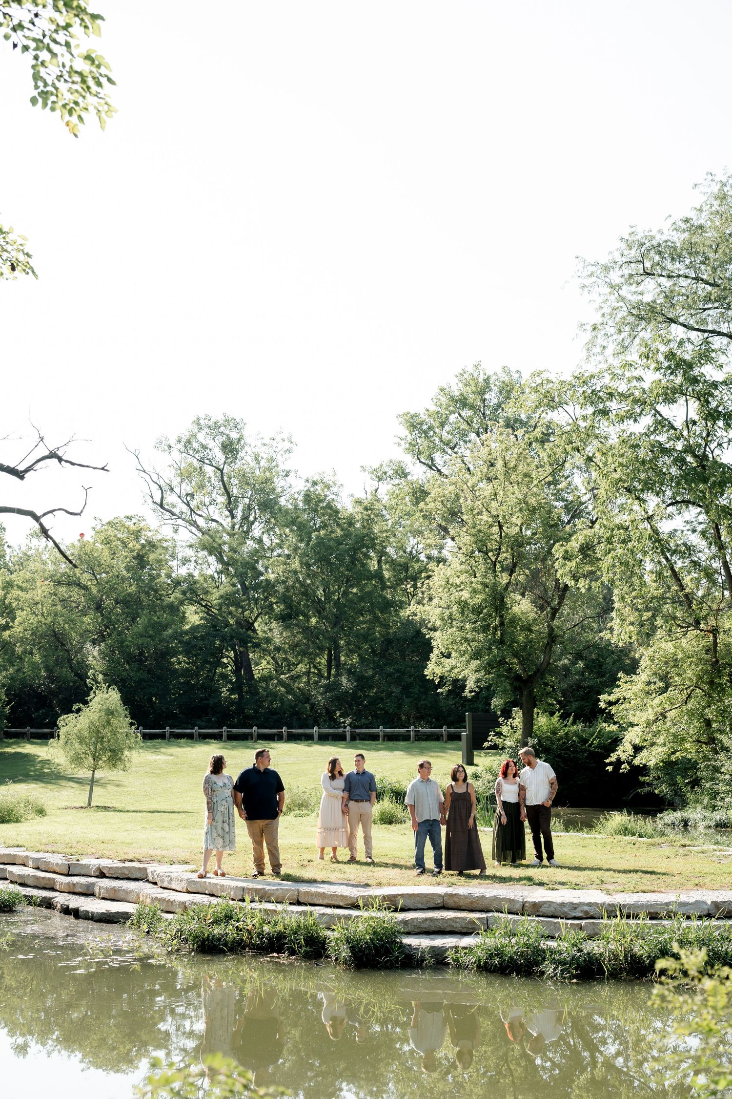 Group of people gathered on stone steps by a reflective pond surrounded by lush green trees on a sunny day.