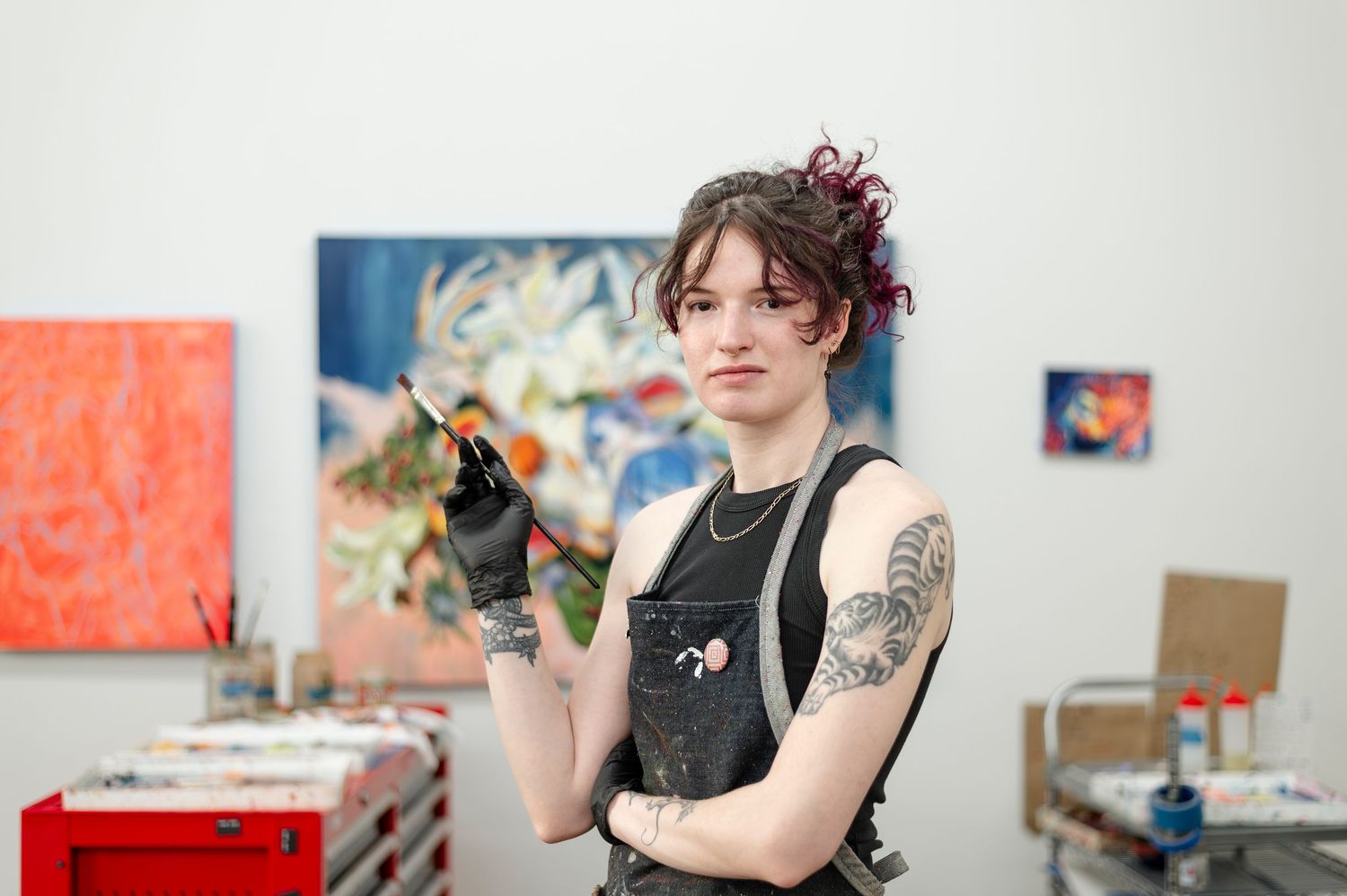 Artist in black tank top with tattoos and pink hair works on painting in bright art studio space.