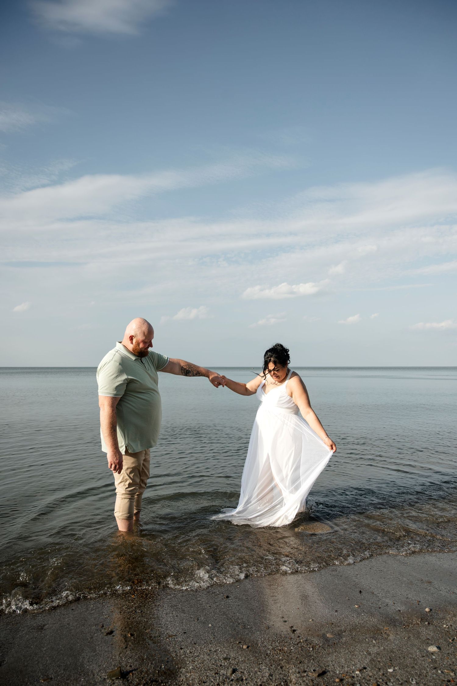 Couple in wedding attire wading in ocean water, bride in white flowing gown holding hands with groom on sunny beach.