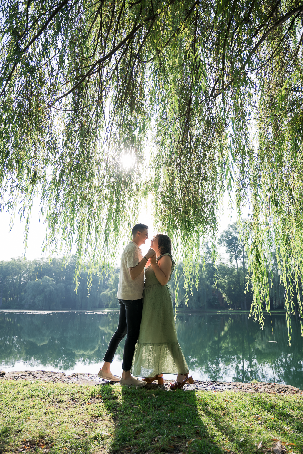 Couple holding hands under a weeping willow tree by a sunlit lake, sharing a romantic moment outdoors.