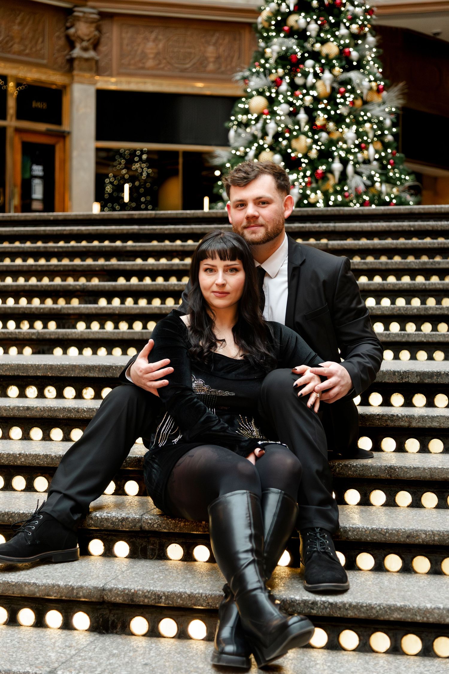 Couple in black outfits sitting on illuminated steps before a decorated Christmas tree in an elegant indoor setting.