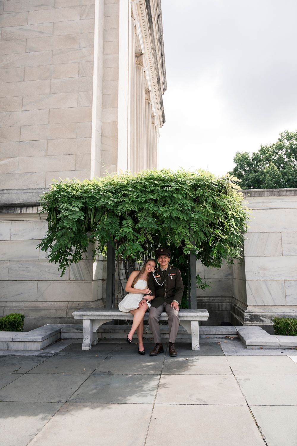 Couple in formal attire sitting on a stone bench under lush ivy beside a grand marble building exterior.