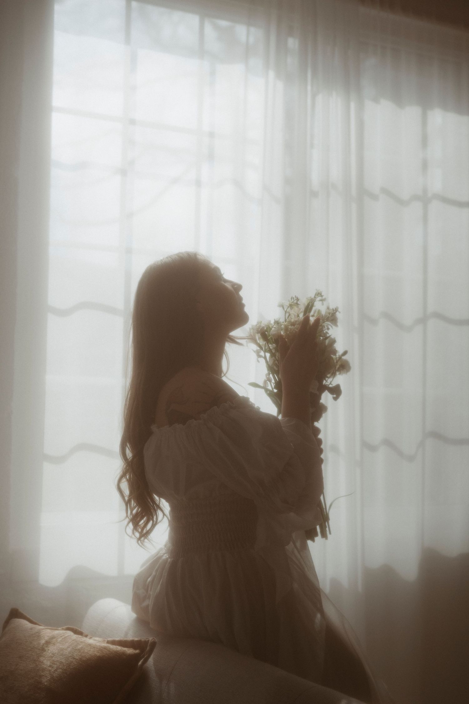 Silhouette of a woman in white off-shoulder dress holding wildflowers by sheer curtained window in soft light.