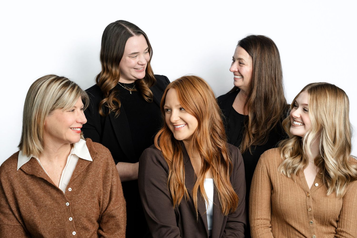 A group of five people wearing brown and neutral toned sweaters laughing together against a white background.