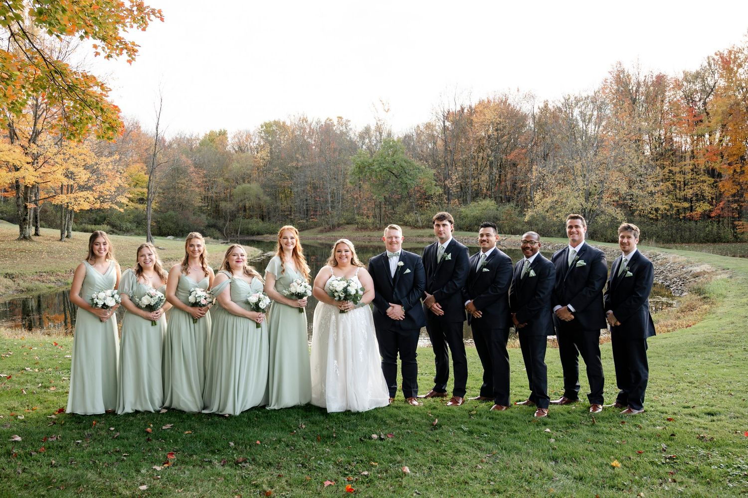 Wedding party in formal attire poses outdoors against autumn trees with golden foliage in the background.