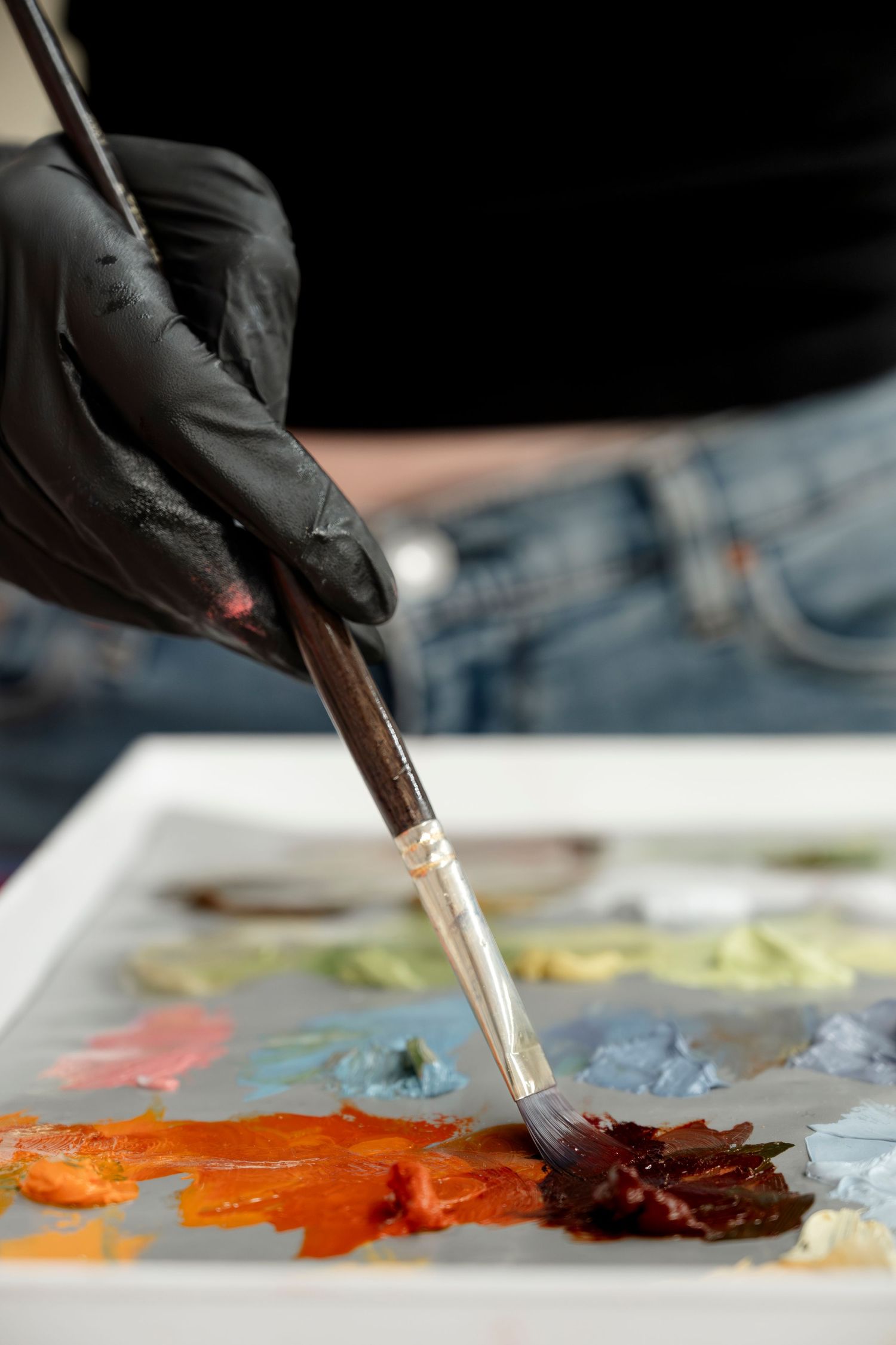 Black gloved hand mixing vibrant orange and brown oil paints on a palette.