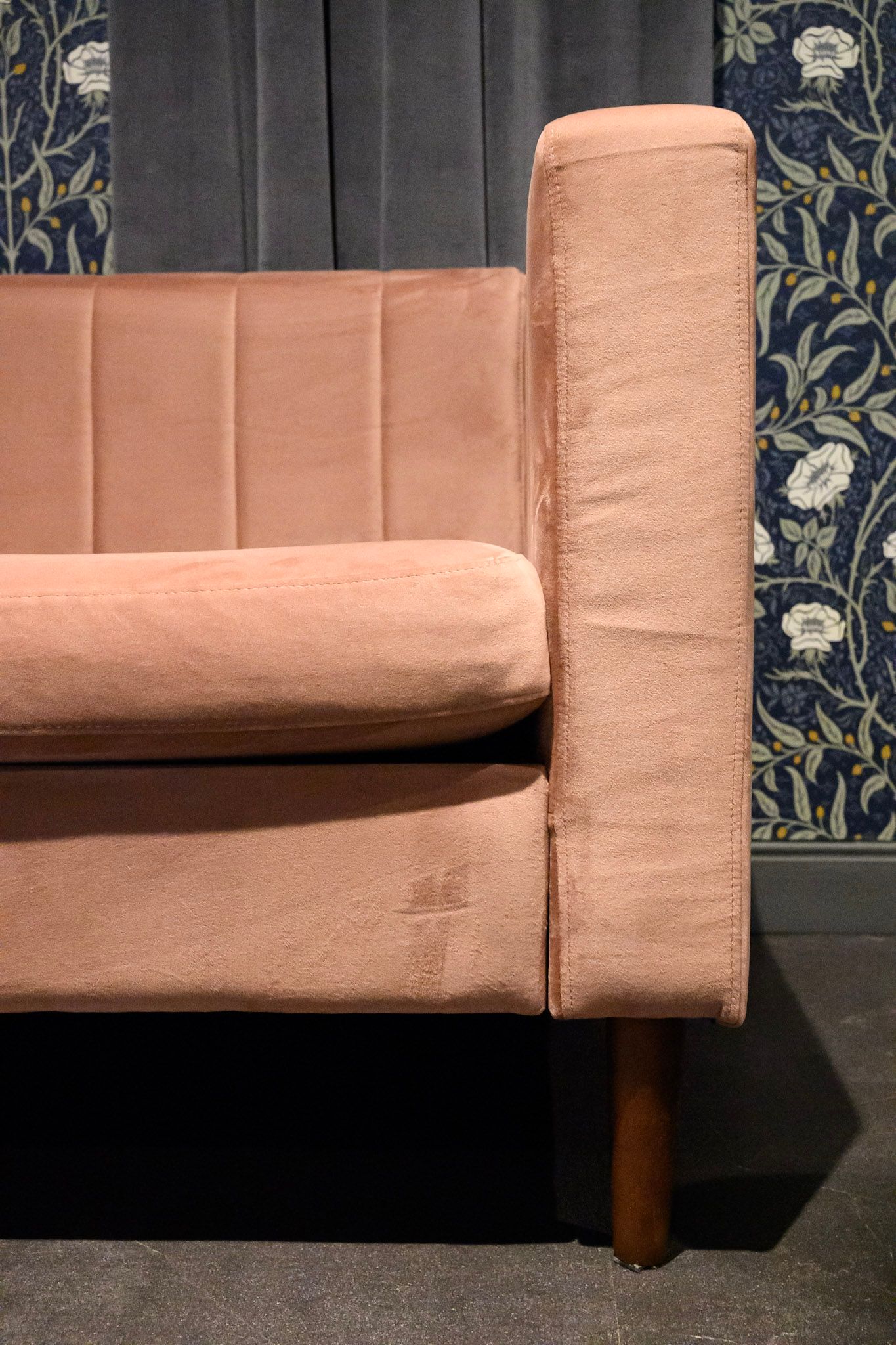 Side view of a peach-colored upholstered sofa with dark wooden legs against a patterned wallpaper background.