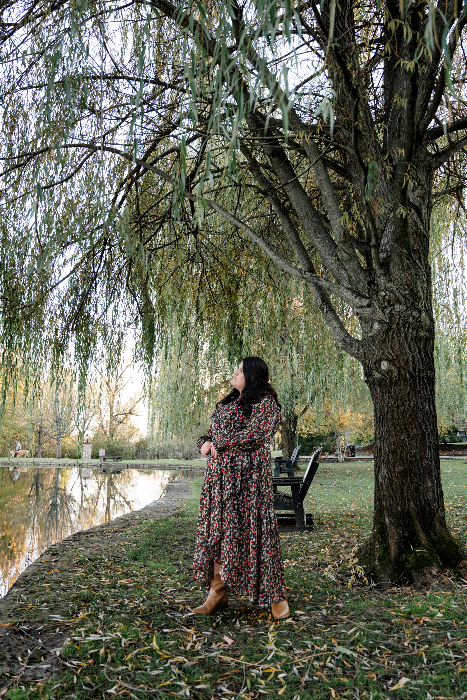 Person in floral dress stands beneath drooping willow tree branches by tranquil pond in autumn setting.