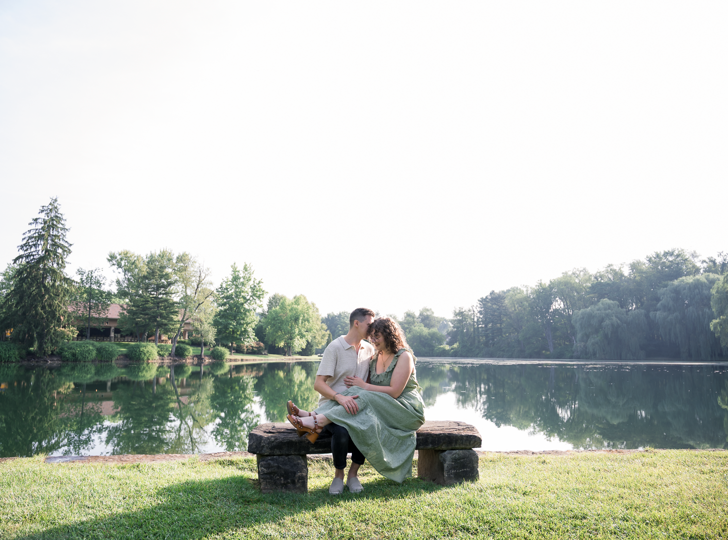 Couple sitting intimately on a bench by a serene lake surrounded by lush green trees.