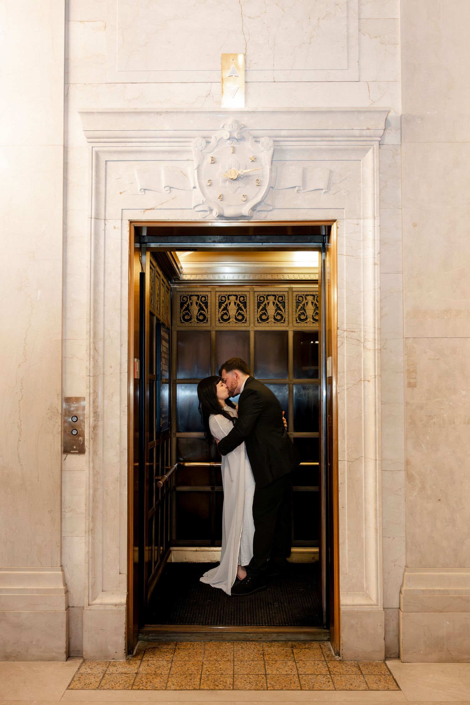Bride and groom sharing a romantic kiss inside a vintage gold elevator at their wedding venue.