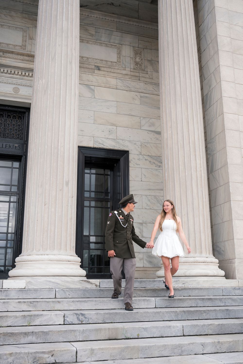Military groom and bride in white dress holding hands on steps of grand building with columns.