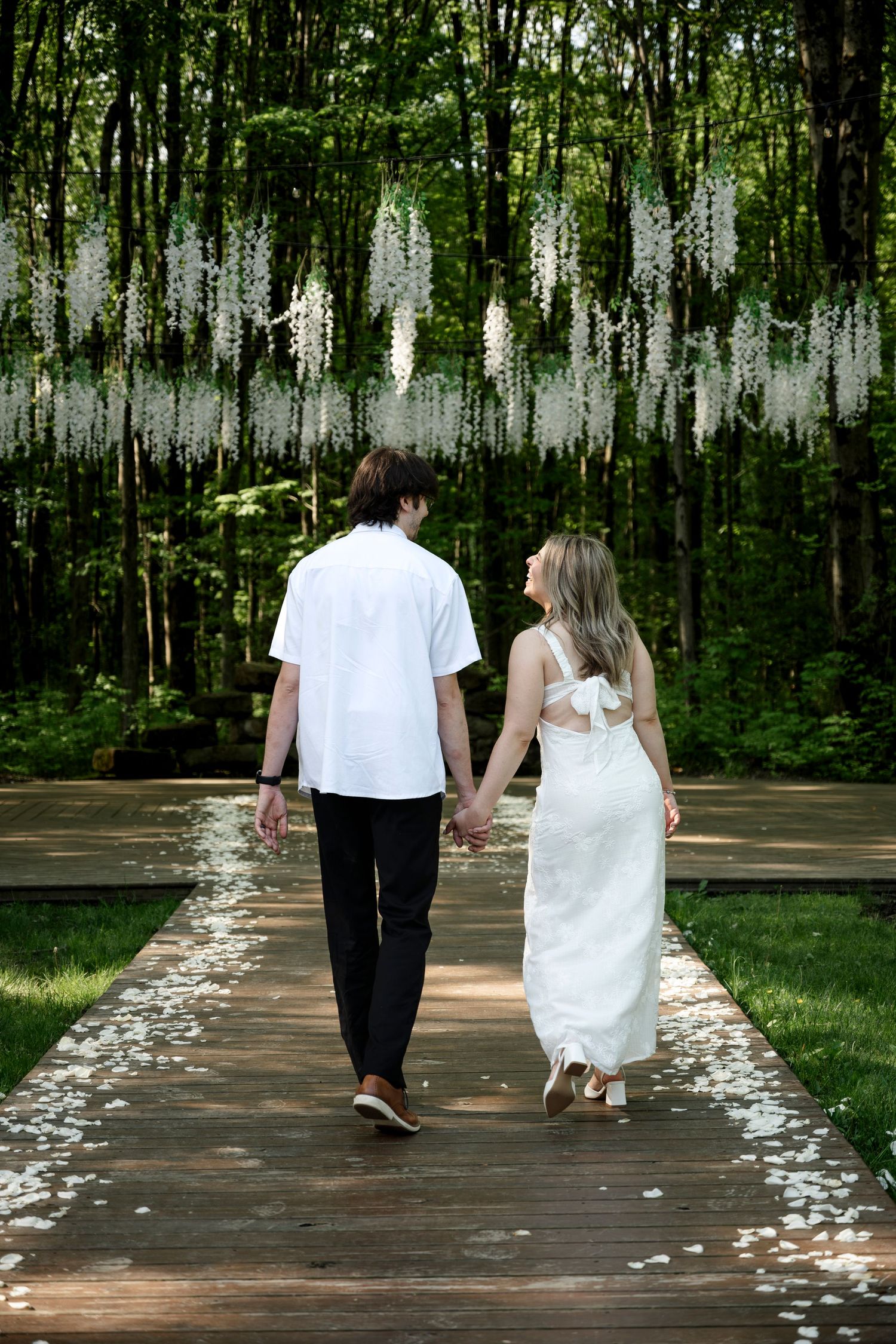 Couple holding hands walking down a petal-lined wooden path beneath hanging white wisteria in a lush forest.