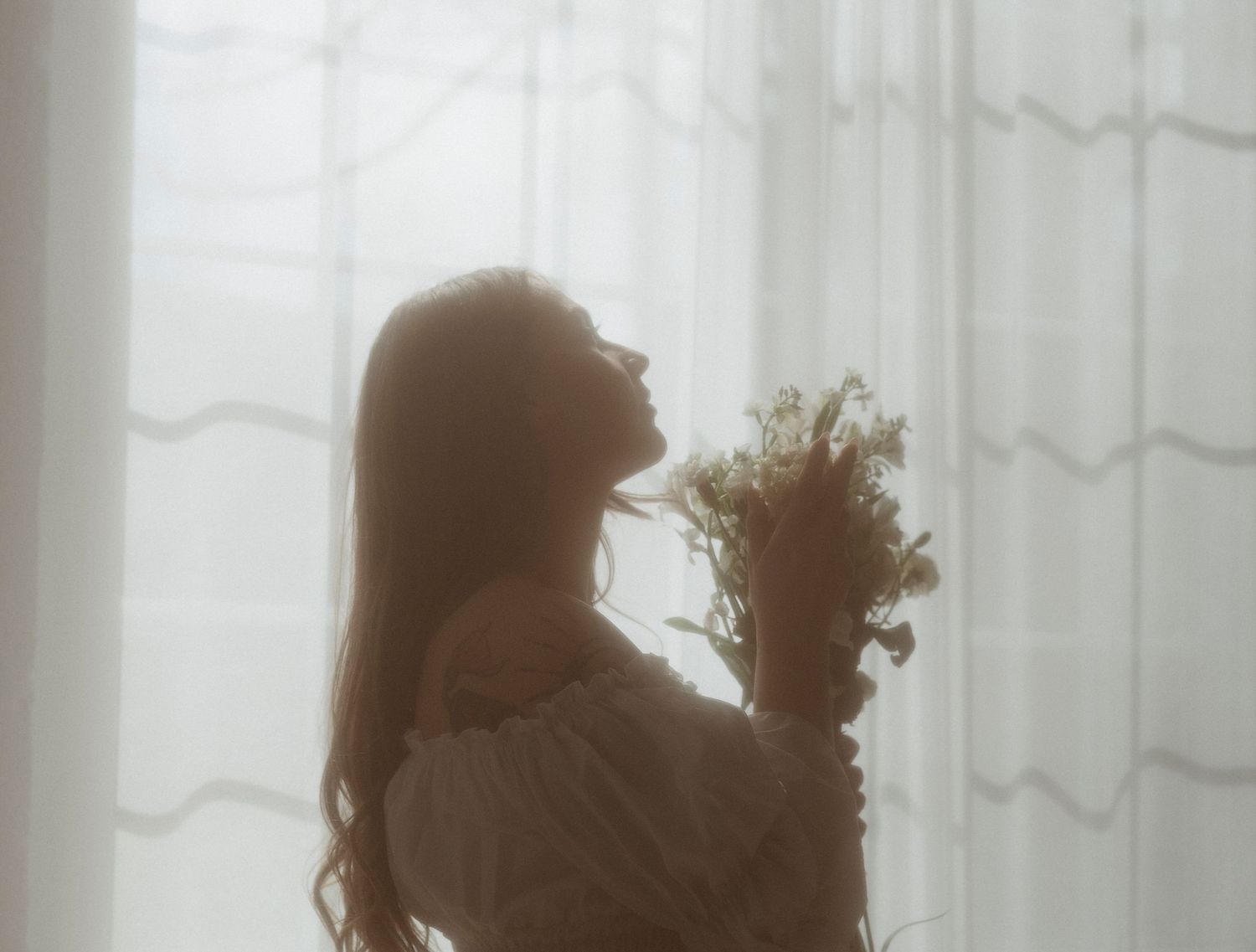 Silhouette of a woman holding a wildflower bouquet by sheer curtains in soft, dreamy backlit light.