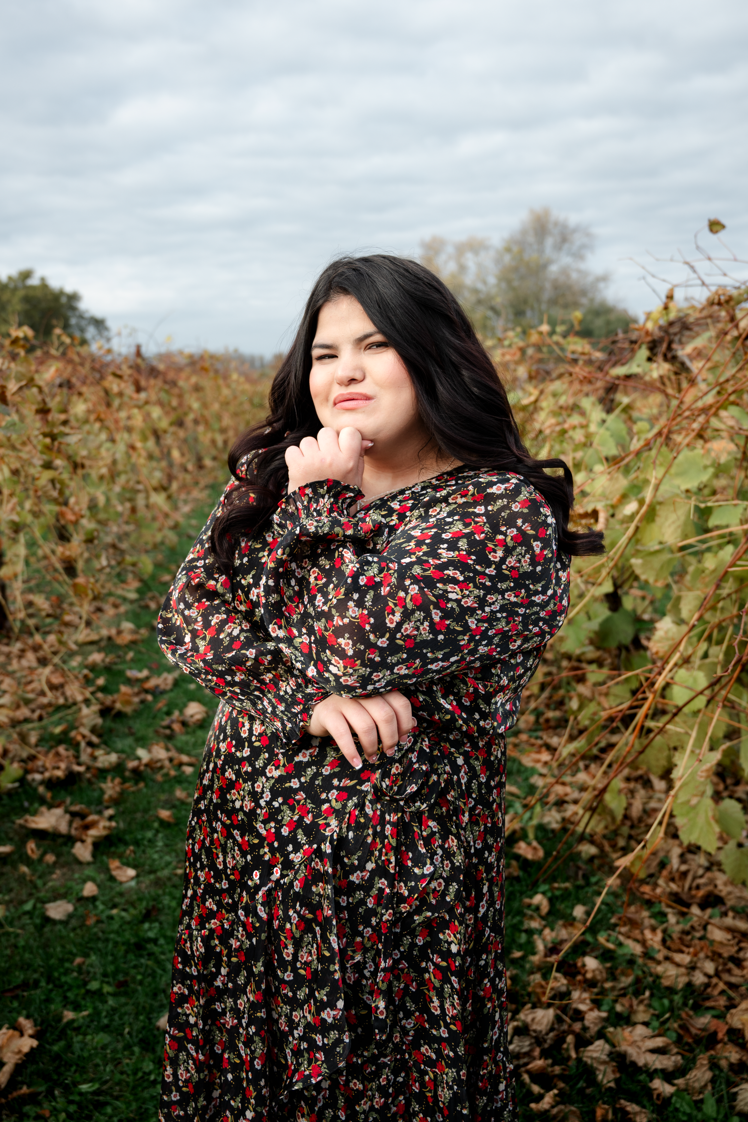 A figure in a floral dress poses expressively against a backdrop of autumn foliage in a natural outdoor setting.
