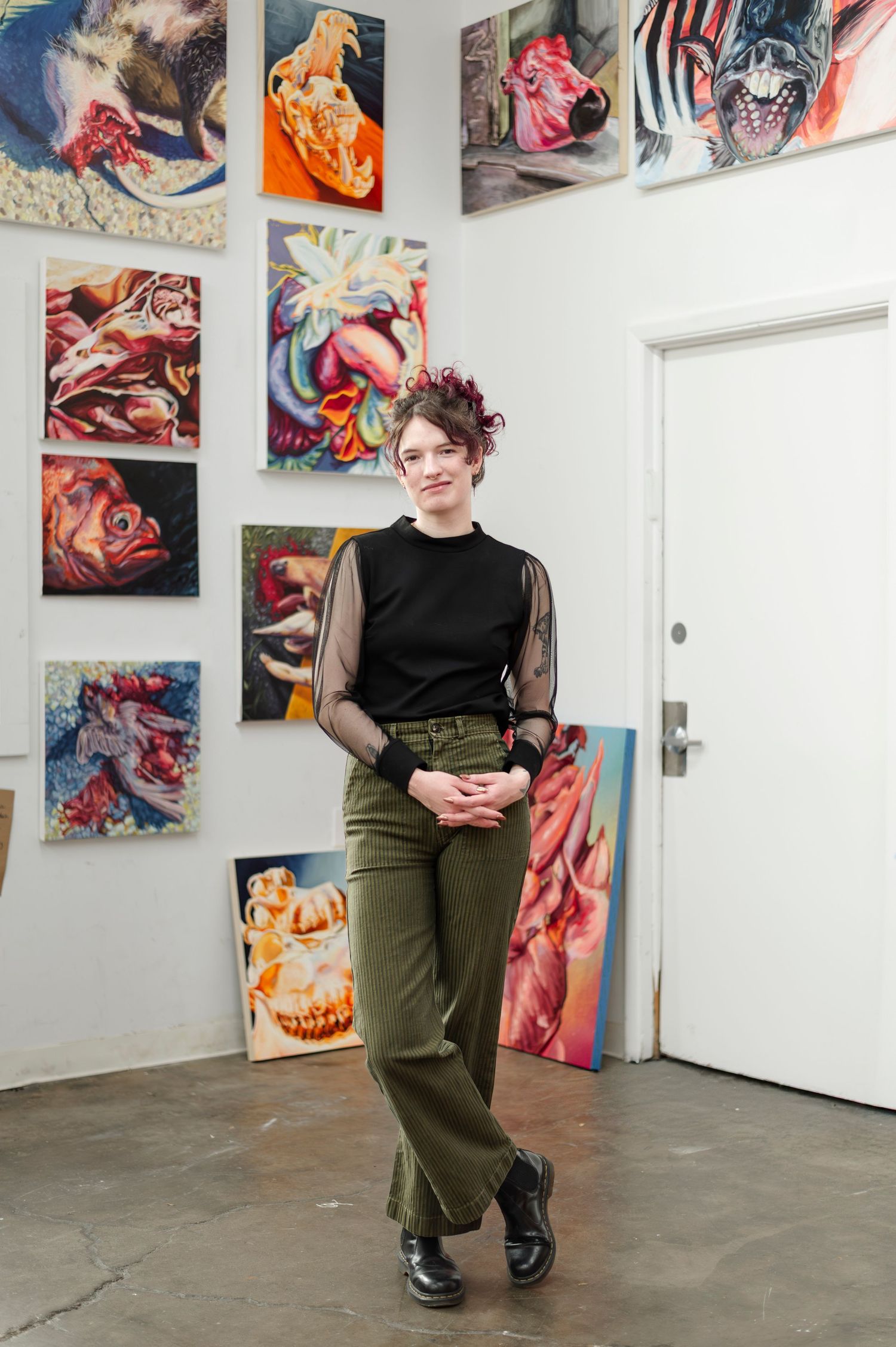 Artist stands in gallery space surrounded by vibrant abstract paintings displayed on white walls.
