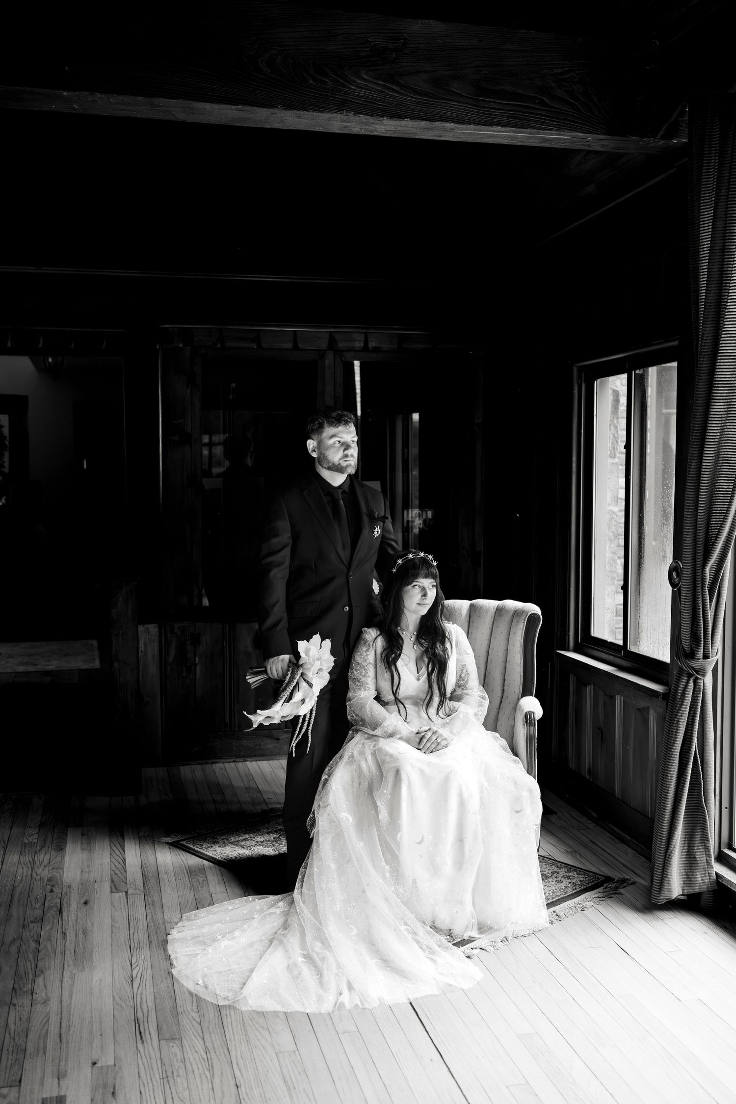 Black and white wedding portrait of bride in lace gown seated while groom stands beside her in dark rustic interior.