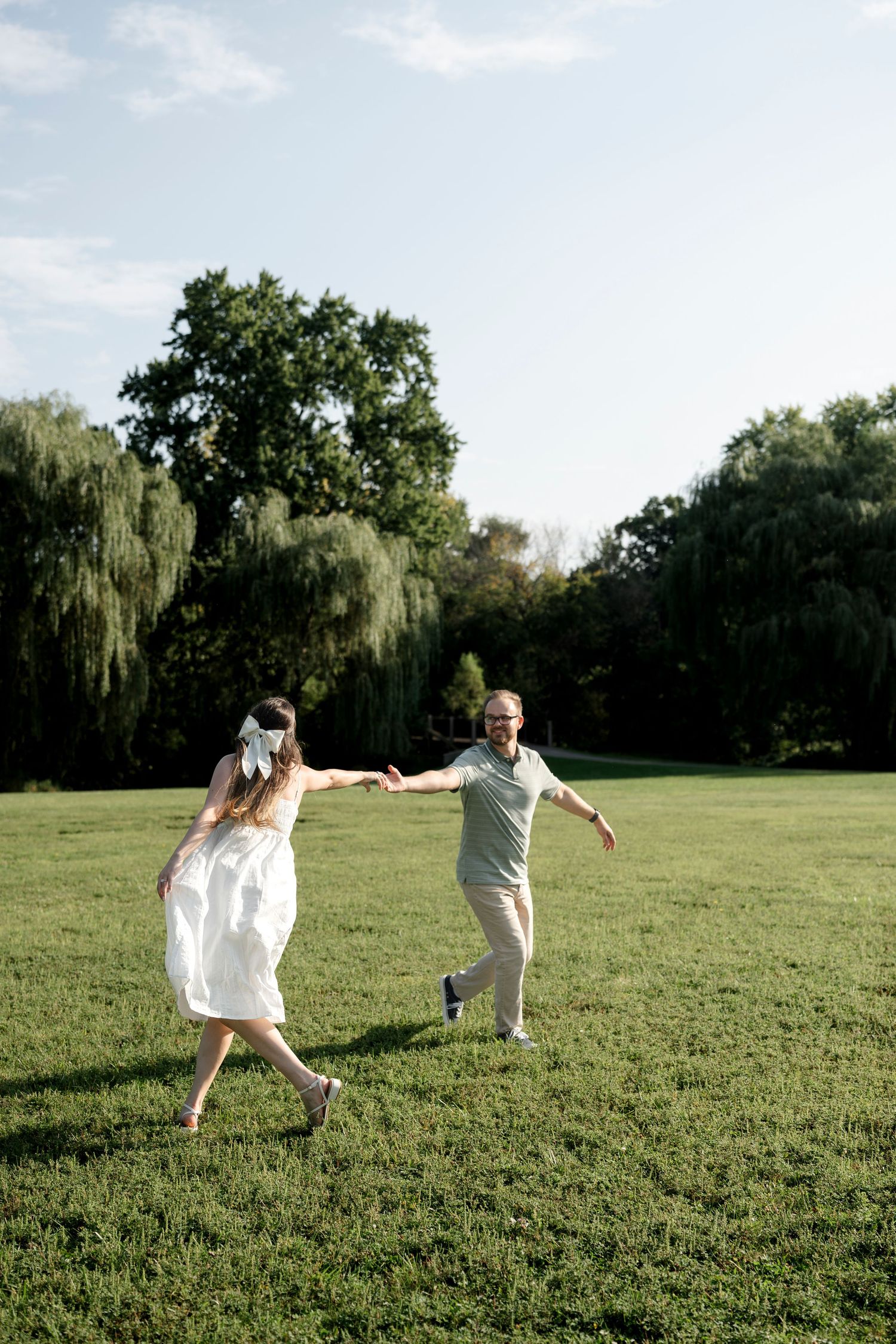 A joyful couple holds hands and dances on a lush green lawn surrounded by trees on a sunny day.