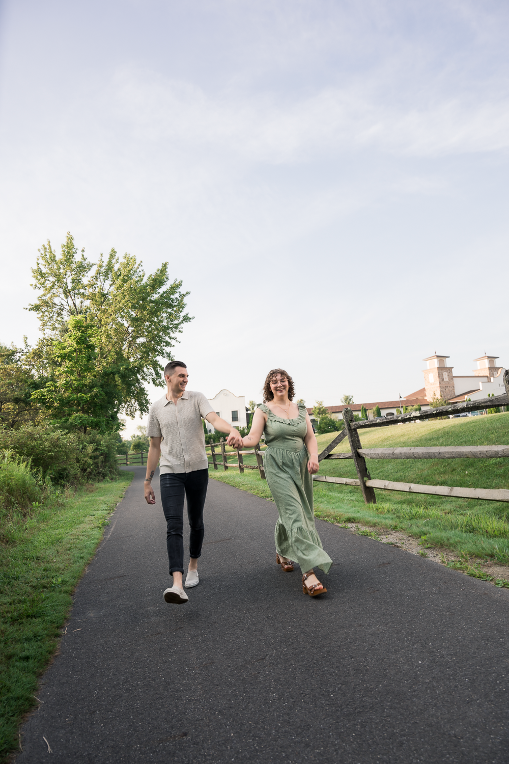 Couple holding hands walking on a paved path outdoors near a wooden fence and green trees on a cloudy day.