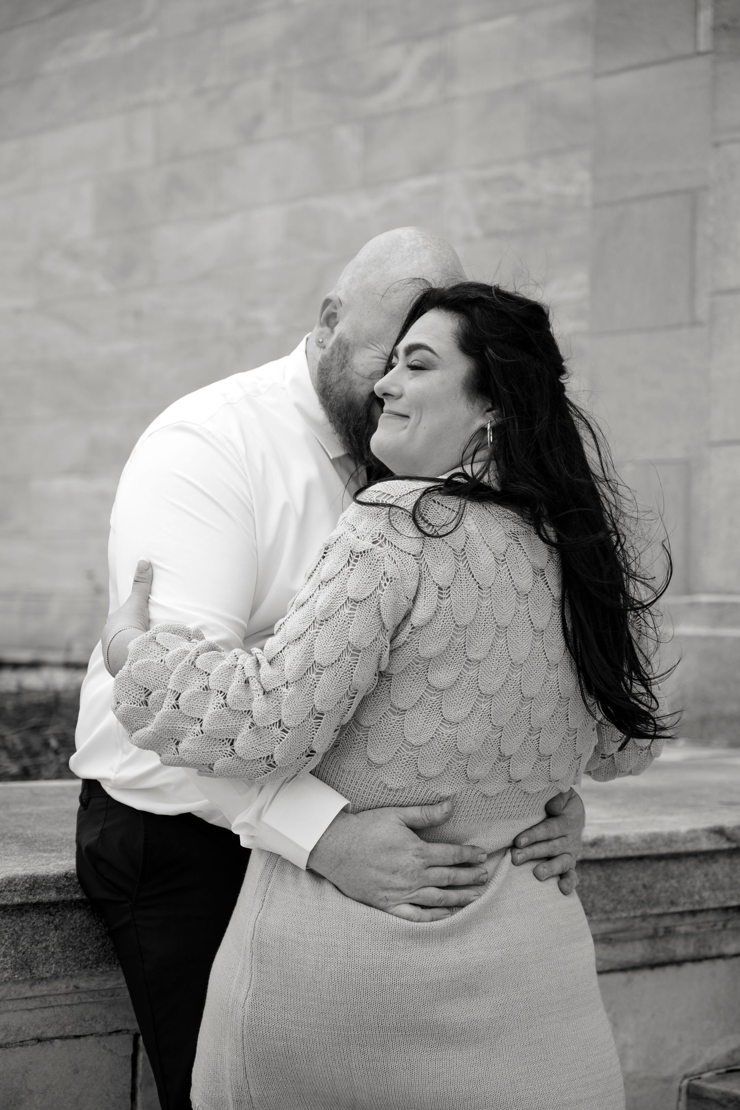 Black and white photo of a couple embracing and kissing near a stone wall, woman smiling in a lace dress.