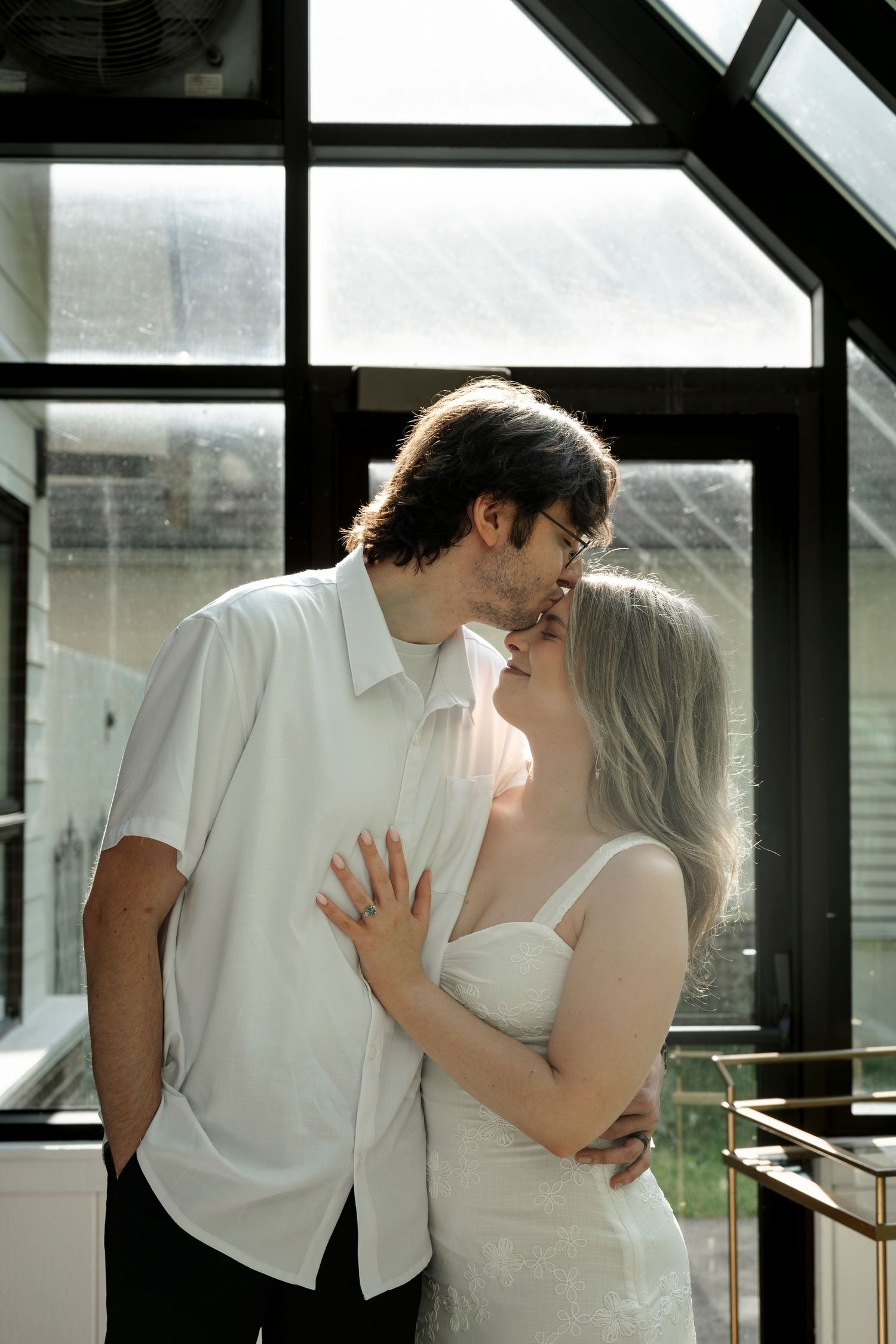 A couple shares a romantic almost-kiss moment, woman in white dress, man in white shirt, near large windows.
