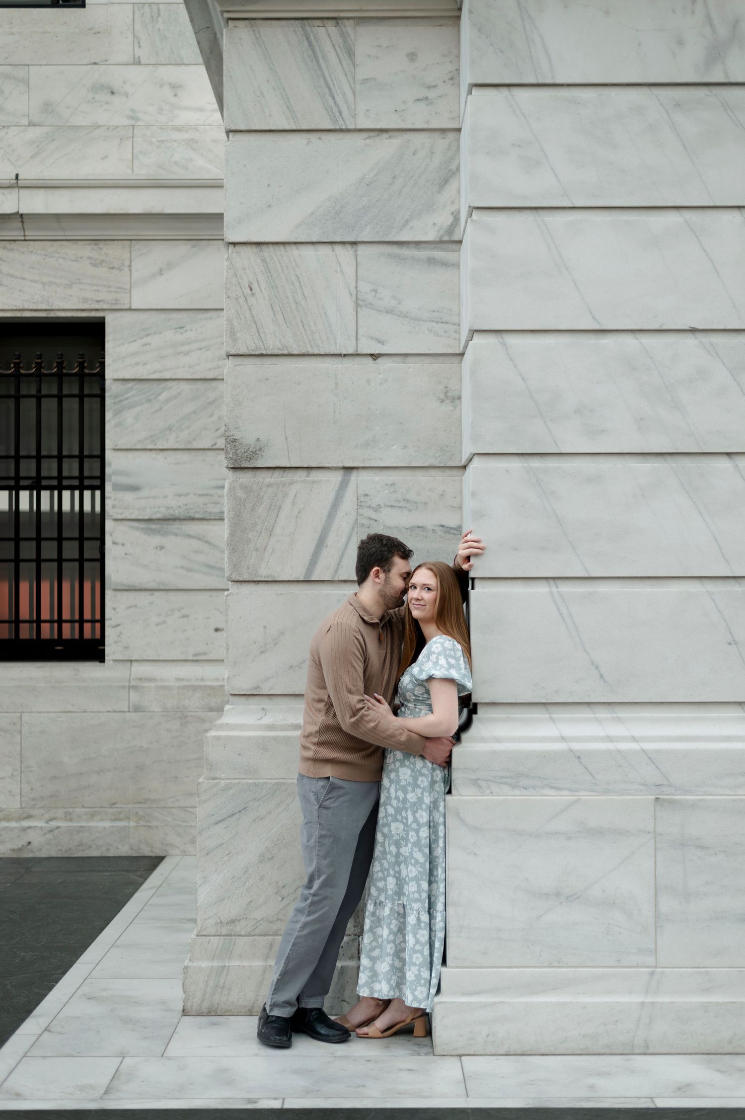 Couple embracing romantically against a marble building exterior, woman in floral dress and man in brown sweater.