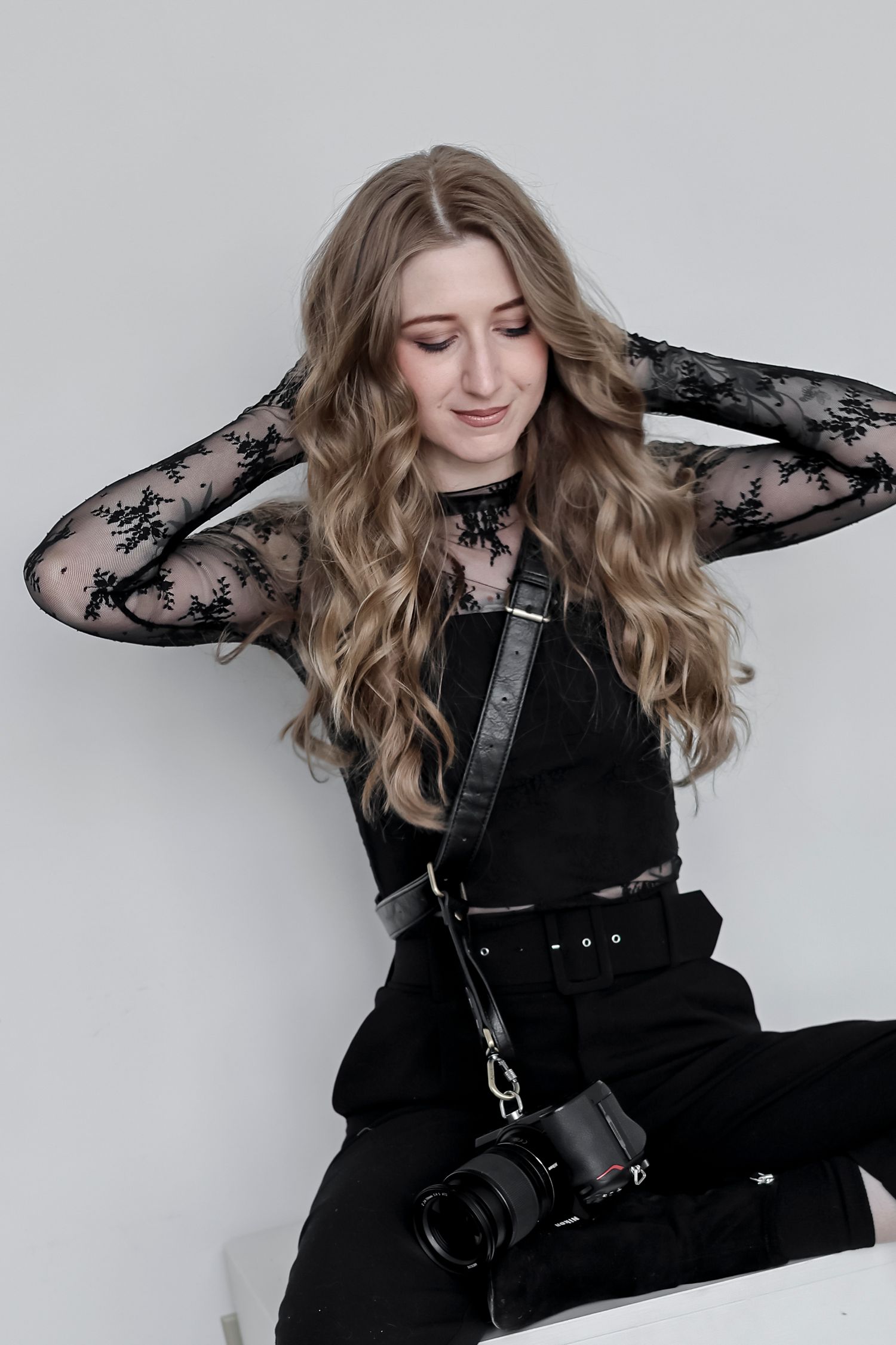 A photographer wearing a black lace top and overalls poses with a camera against a light gray background.