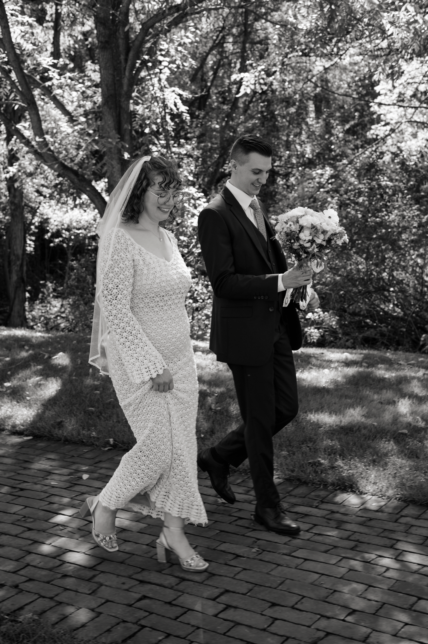 A couple in formal attire walks together on a brick pathway surrounded by dappled sunlight and trees.
