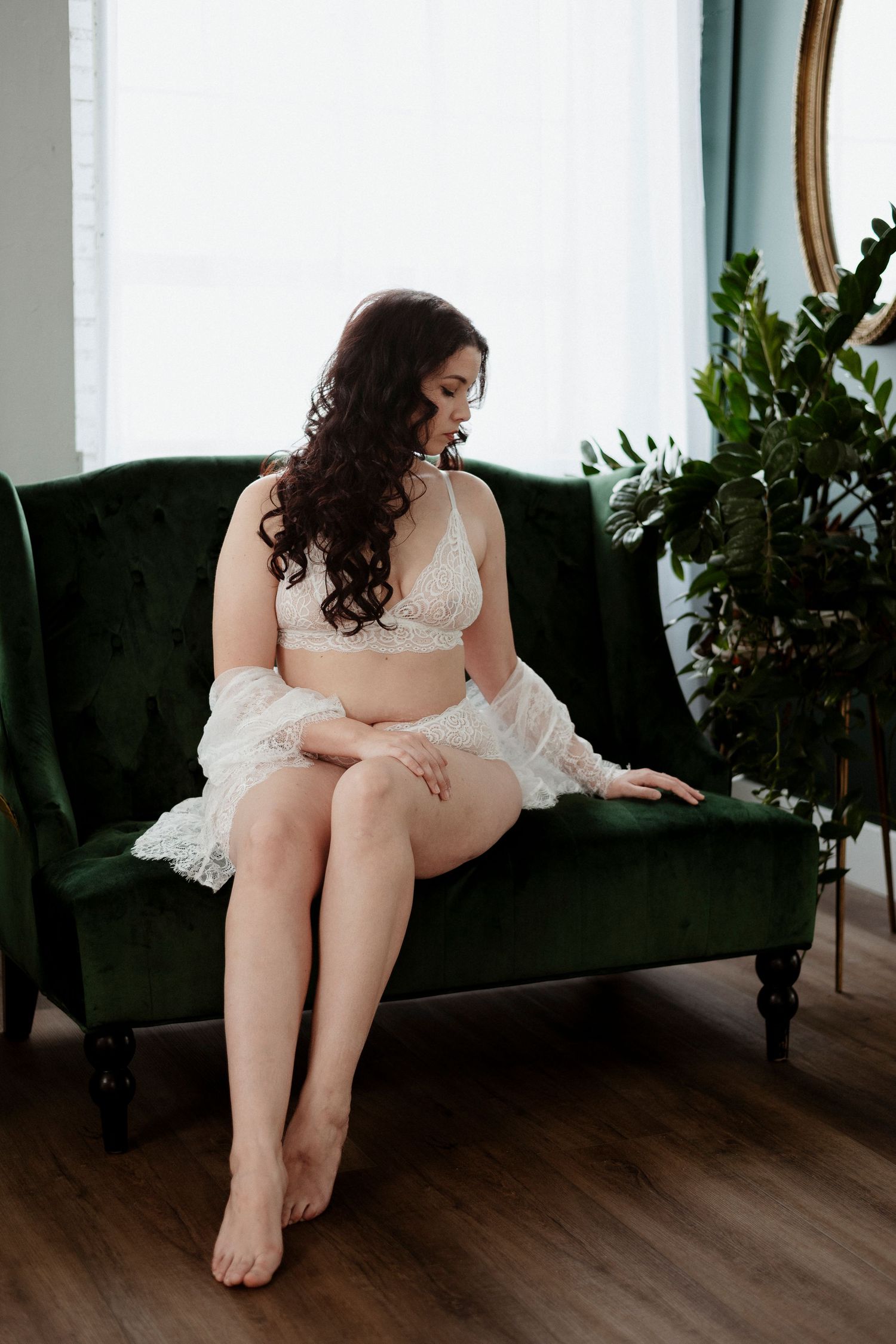 A woman in white lingerie reclines on a dark velvet couch near potted plants in a sunlit room.