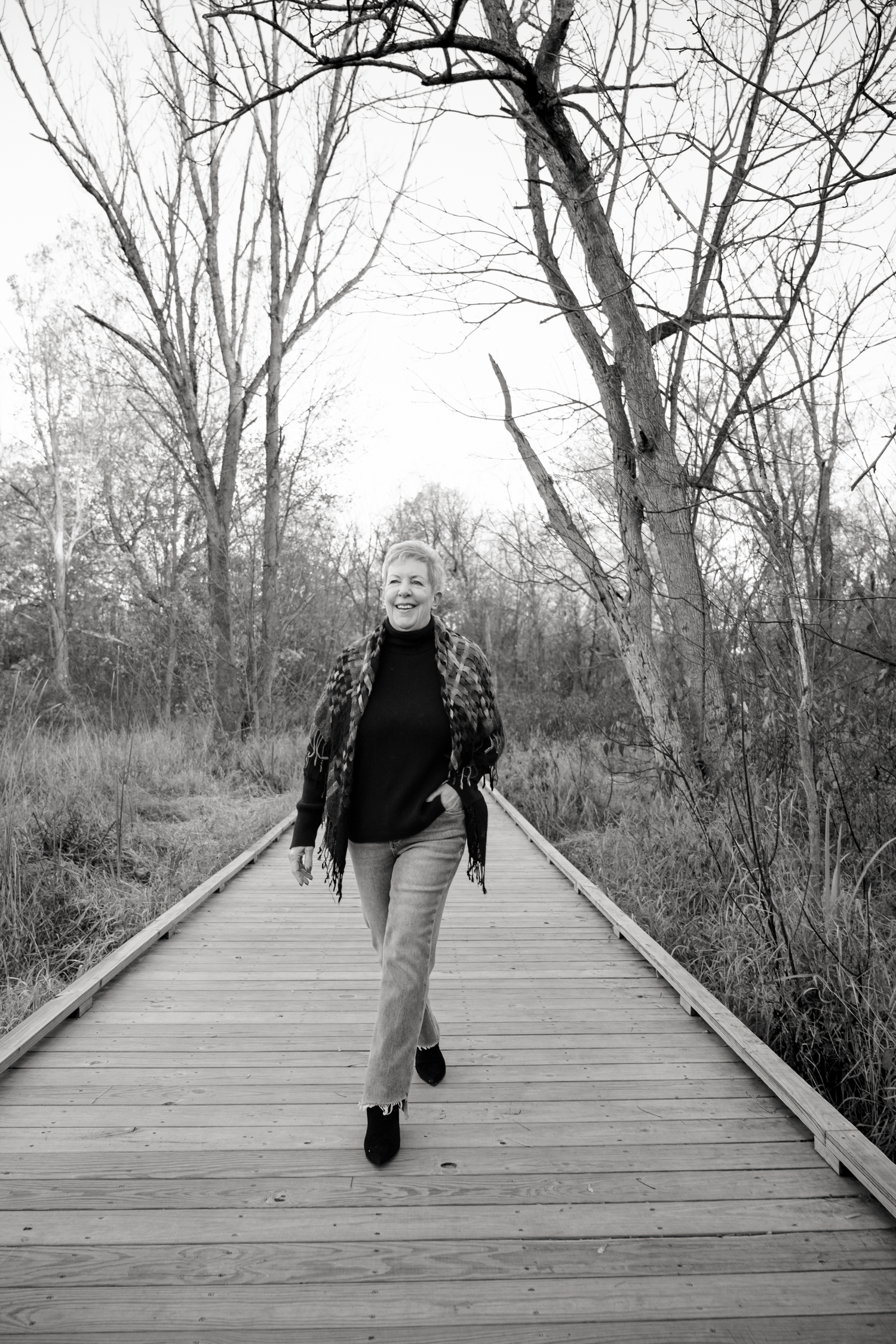 Black and white photo of a person walking away on a wooden boardwalk through bare trees.