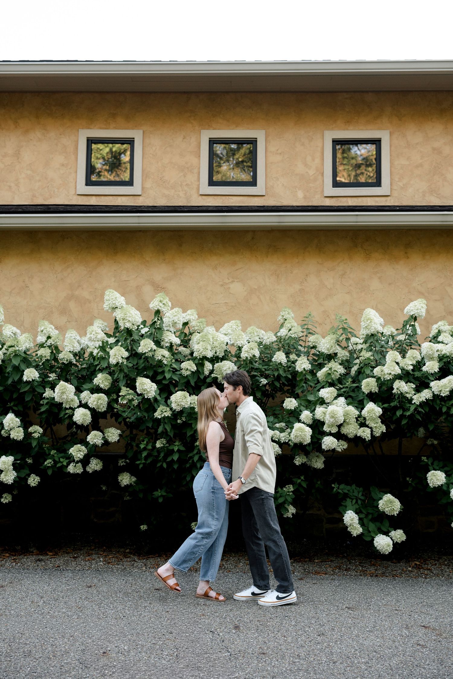 Couple holding hands and almost kissing in front of white hydrangea bushes and a yellow stucco building.
