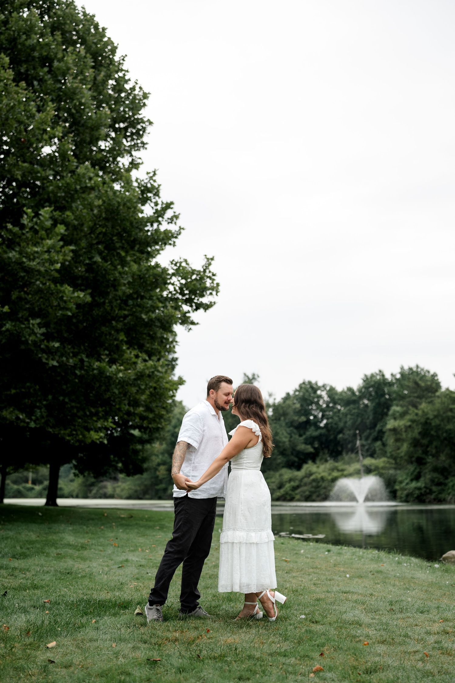 Couple sharing a romantic kiss in a lush green park with a pond and fountain in the background.