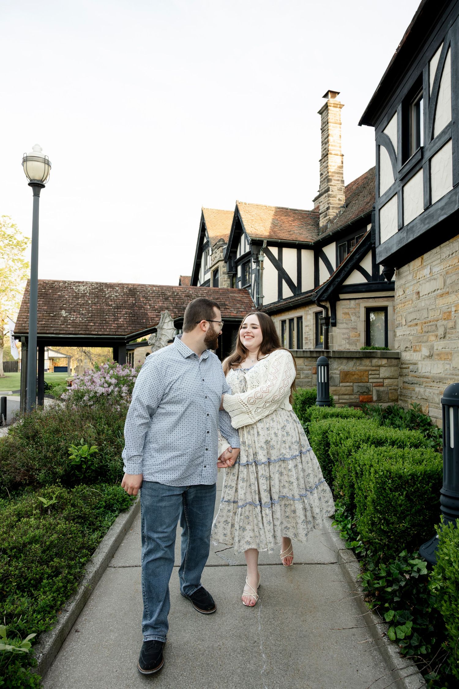 Couple walks hand-in-hand on a garden path beside a Tudor-style building, smiling at each other on a spring day.