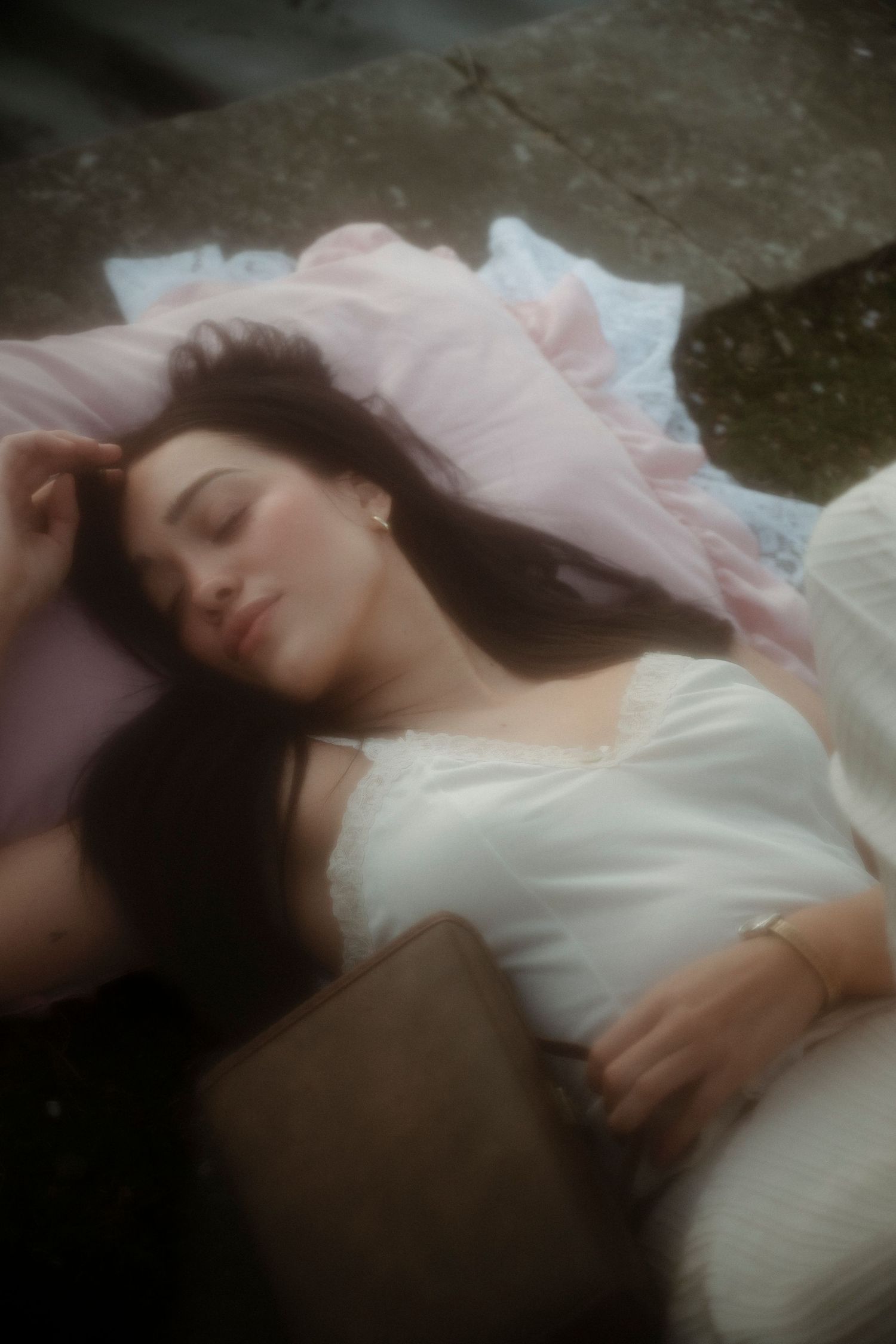 A young woman with long dark hair rests peacefully on pink pillows outdoors, wearing a white lace top.