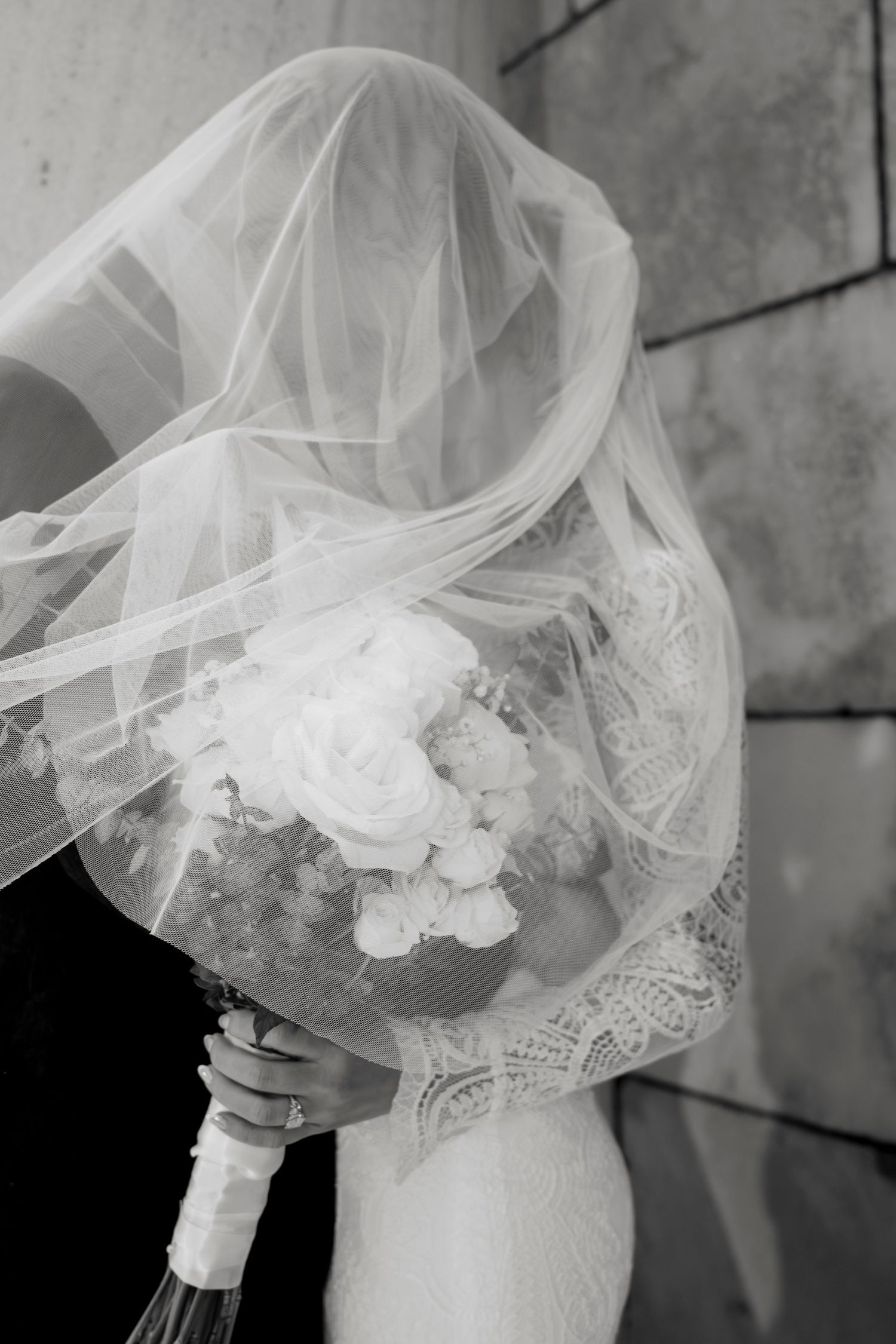 A black and white image of a bride holding a bouquet of flowers, with her face obscured by a flowing veil.