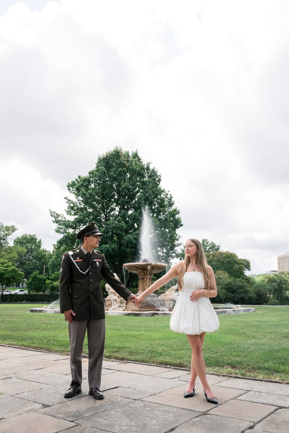 Military man in uniform holds hands with woman in white dress by a fountain in a lush green park.
