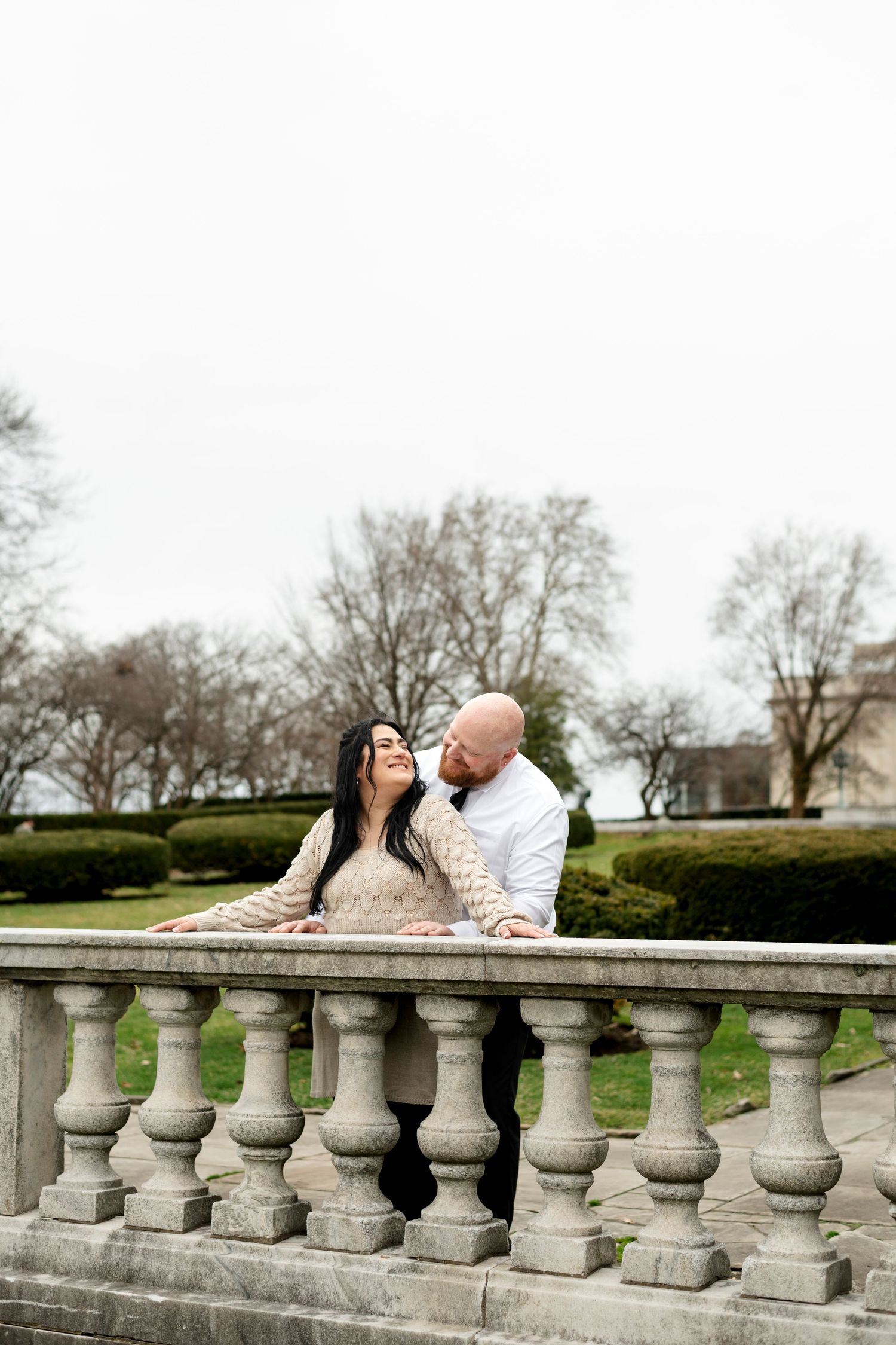 Couple shares romantic moment leaning on stone balustrade in park, woman in beige dress smiling up at bald man in white shirt.