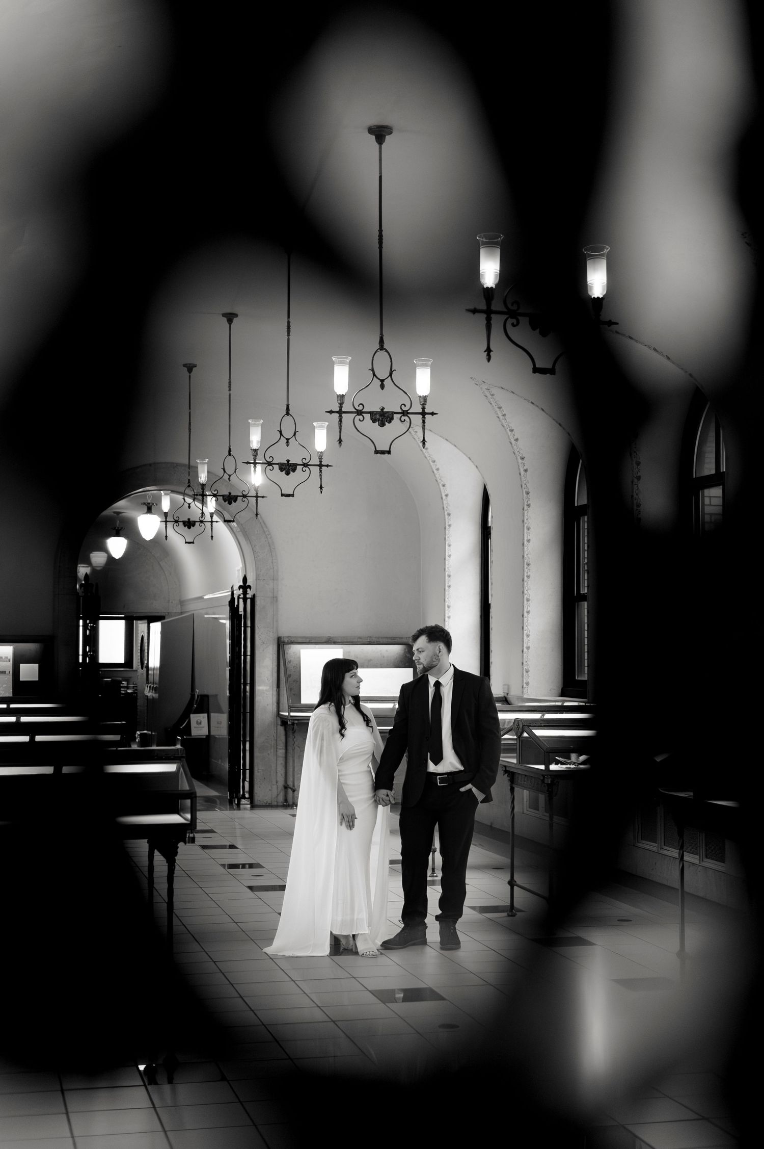 Black and white photo of a couple in wedding attire standing in an elegant hall with chandeliers.
