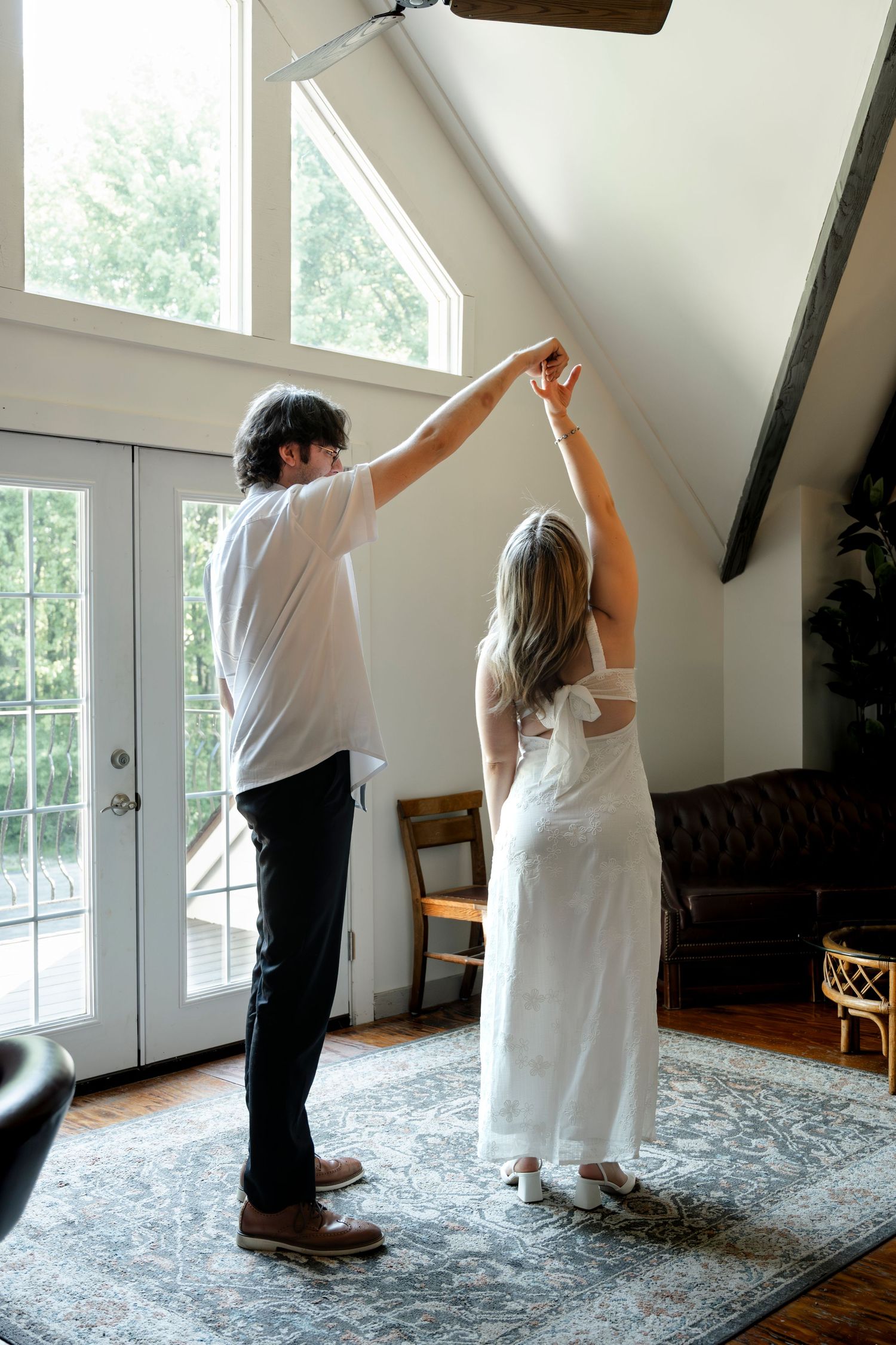 Couple dancing indoors, man in white shirt spinning woman in white dress on a patterned rug in a bright living room.