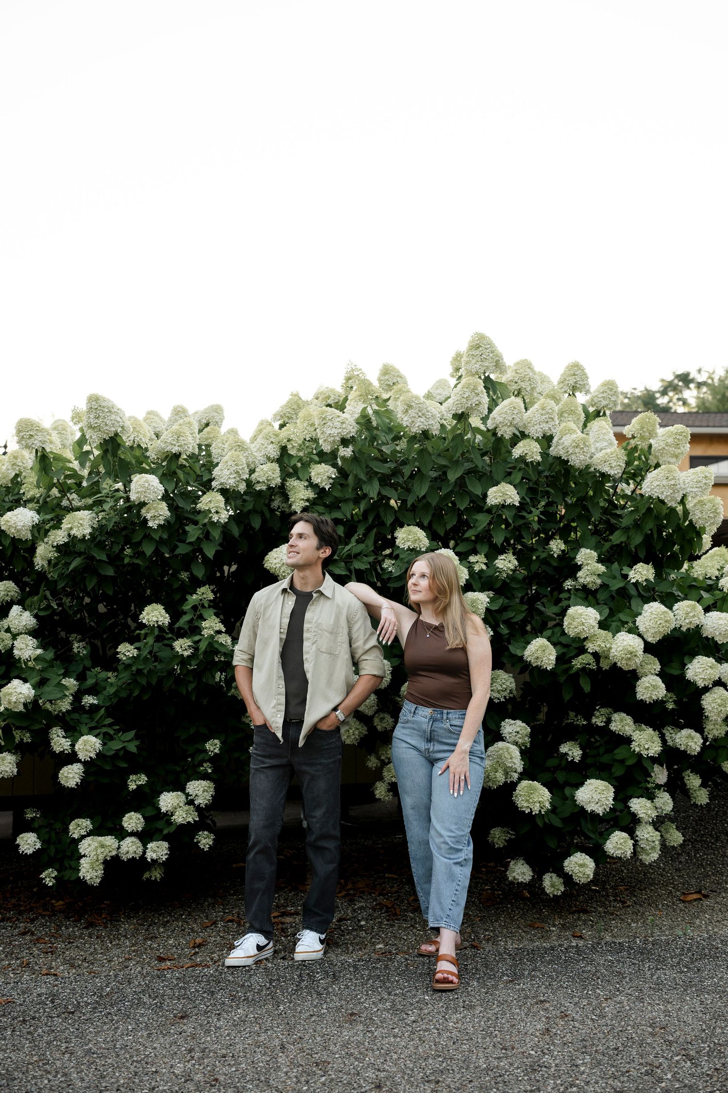 A couple stands in front of lush white hydrangea bushes, the woman resting her arm on the man's shoulder.
